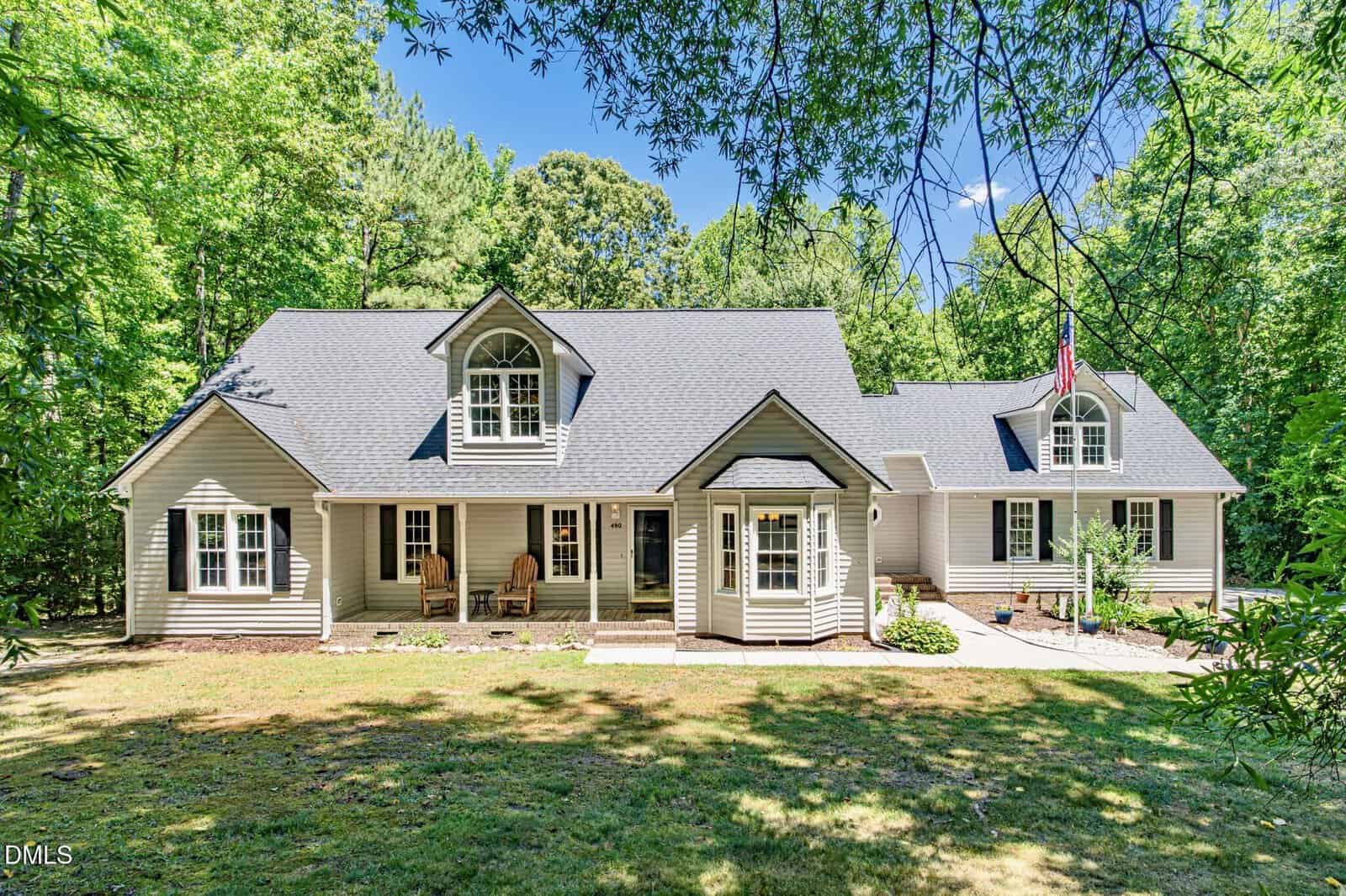 A charming two-story house at 490 Huntsburg Drive in Louisburg, with light-colored siding, dormer windows, a covered front porch with chairs, a bay window, and an American flag. Surrounded by trees and grass on a sunny day.