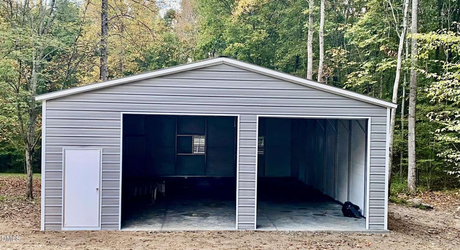 A gray metal garage with two large open bays and a side door, surrounded by trees with green and yellow leaves in a wooded area.