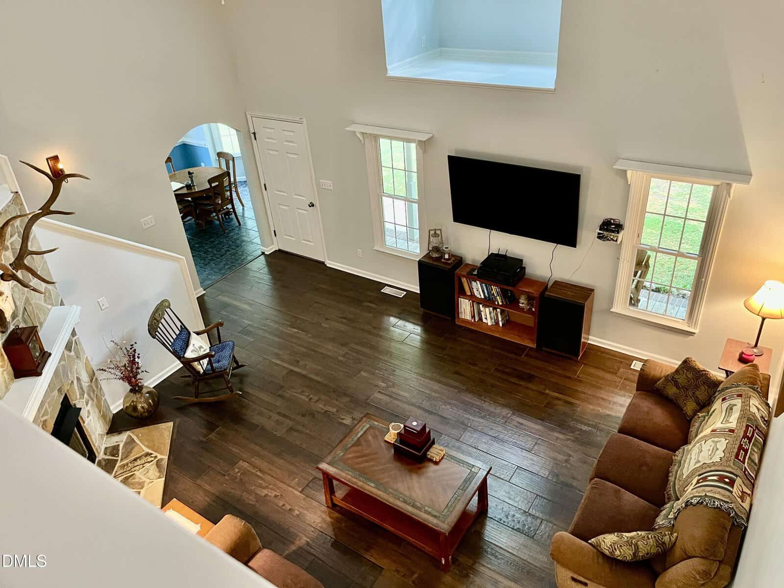 A spacious living room with hardwood floors, a brown sofa, armchair, wall-mounted TV, coffee table, bookshelf, and large windows. Adjacent dining room visible through an arched doorway.