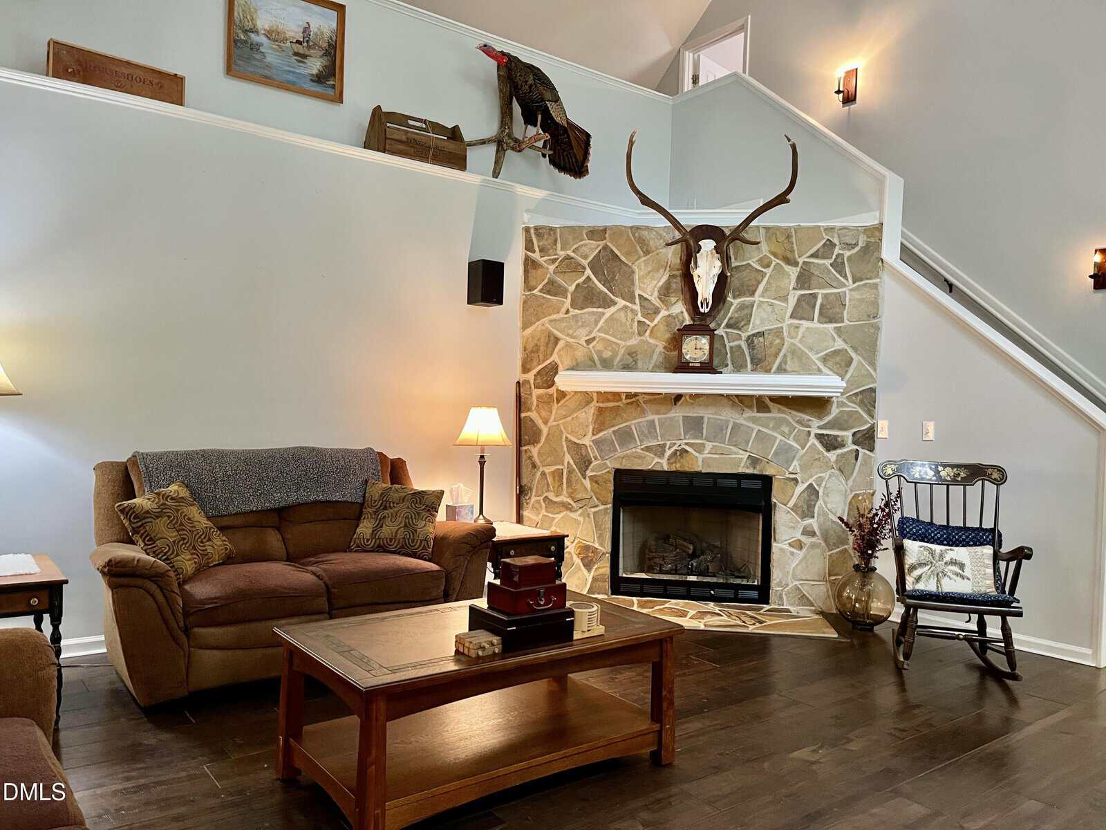 Cozy living room with brown couches, a stone fireplace, mounted animal skull, wooden coffee table, rocking chair, and decorative items on shelves and walls. Stairs lead up along the right side.