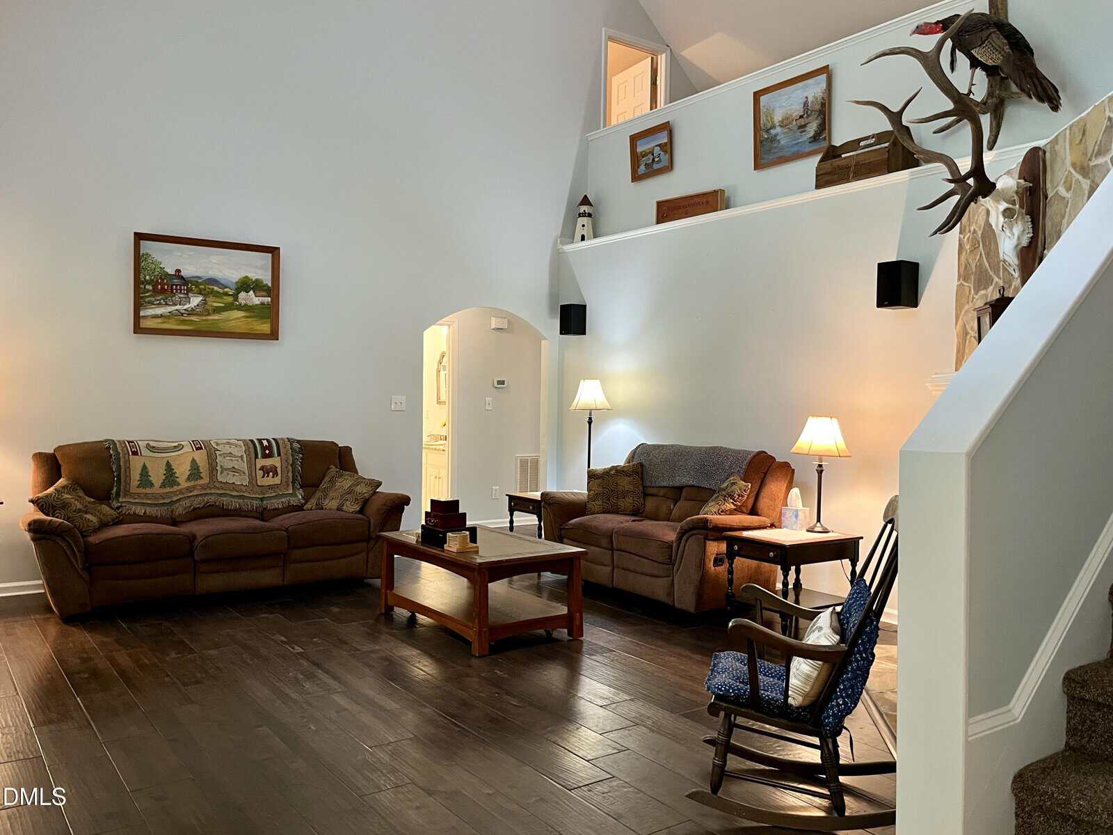 A cozy living room with two brown sofas, a coffee table, lamps, a rocking chair, and wall art. Stairs lead up to a loft area with more decorations, including antlers and framed pictures.