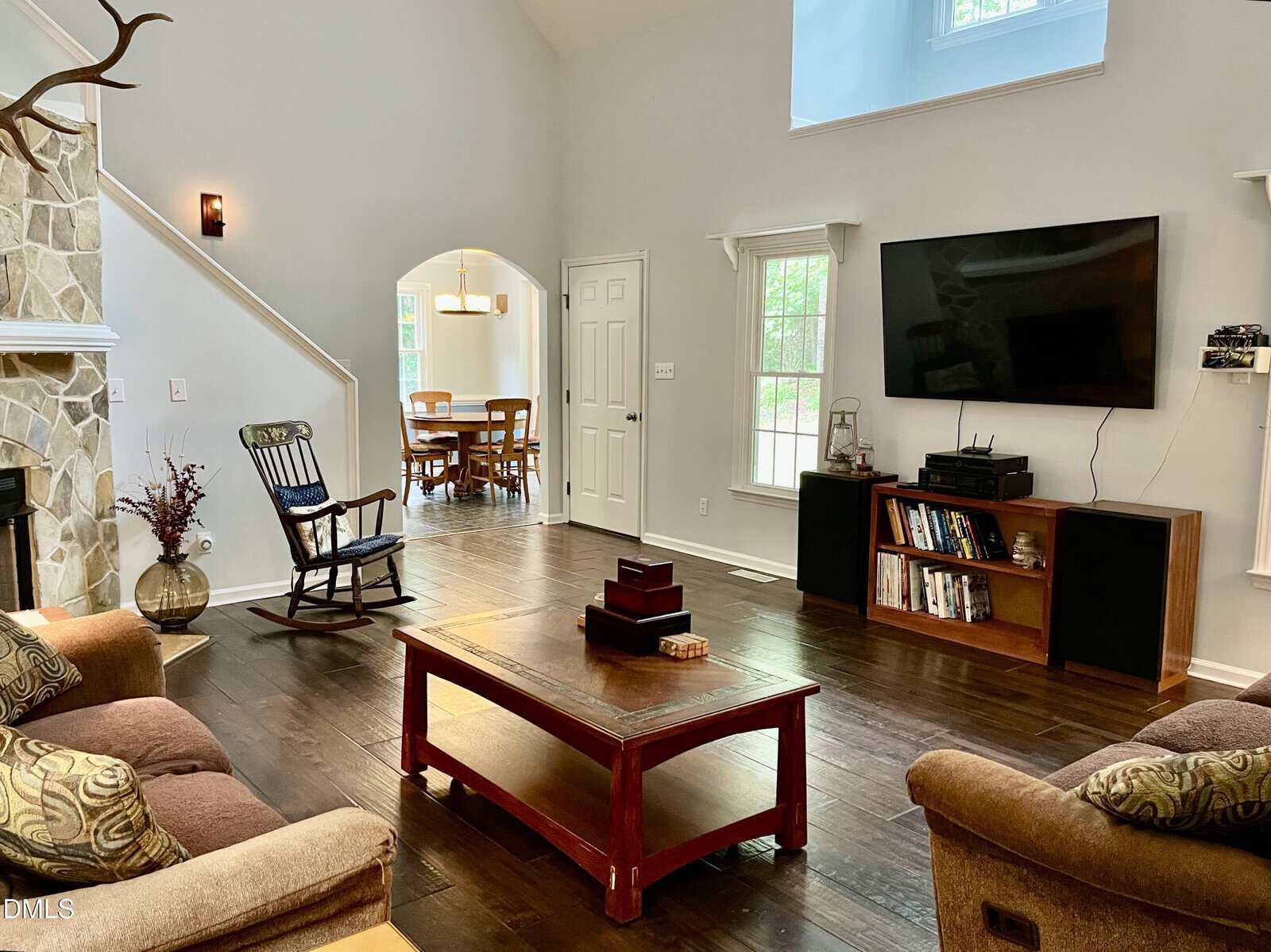 A spacious living room with wooden floors, brown sofas, a stone fireplace, rocking chair, coffee table, bookshelves, and a wall-mounted TV. Large windows and a dining area can be seen in the background.