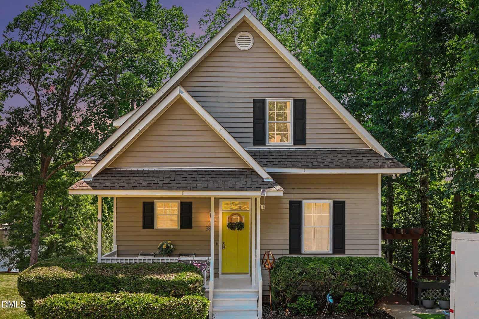A two-story beige house with a steep gable roof, black shutters, and a bright yellow front door. Bushes line the front yard, and trees surround the home, with steps leading up to the porch.
