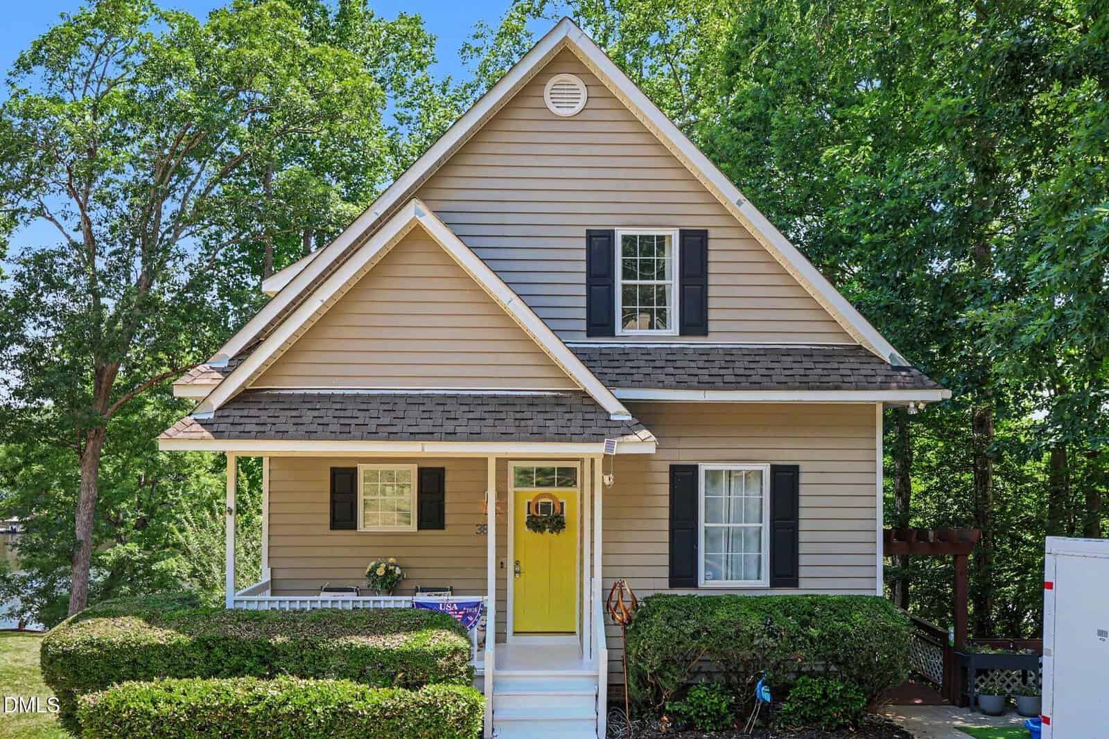 A beige two-story house at 383 Sagamore Drive, Louisburg, with white trim, black shutters, and a bright yellow front door. Green bushes and tall trees surround the small porch with white railings and steps leading to the entrance.