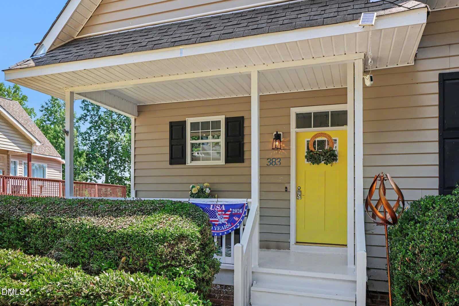A beige house with white trim features a covered front porch, a yellow door with a wreath, a window with black shutters, and a small USA-themed banner draped over the porch railing. Bushes line the walkway.