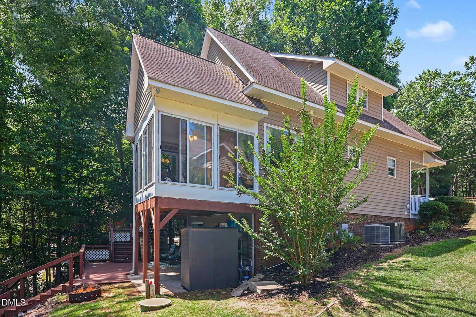 A two-story beige house with a screened-in porch overlooks a wooded yard. The house has a sloped roof, elevated deck area, and the yard is partially shaded by tall trees. Shrubs and landscaping are visible around the house.