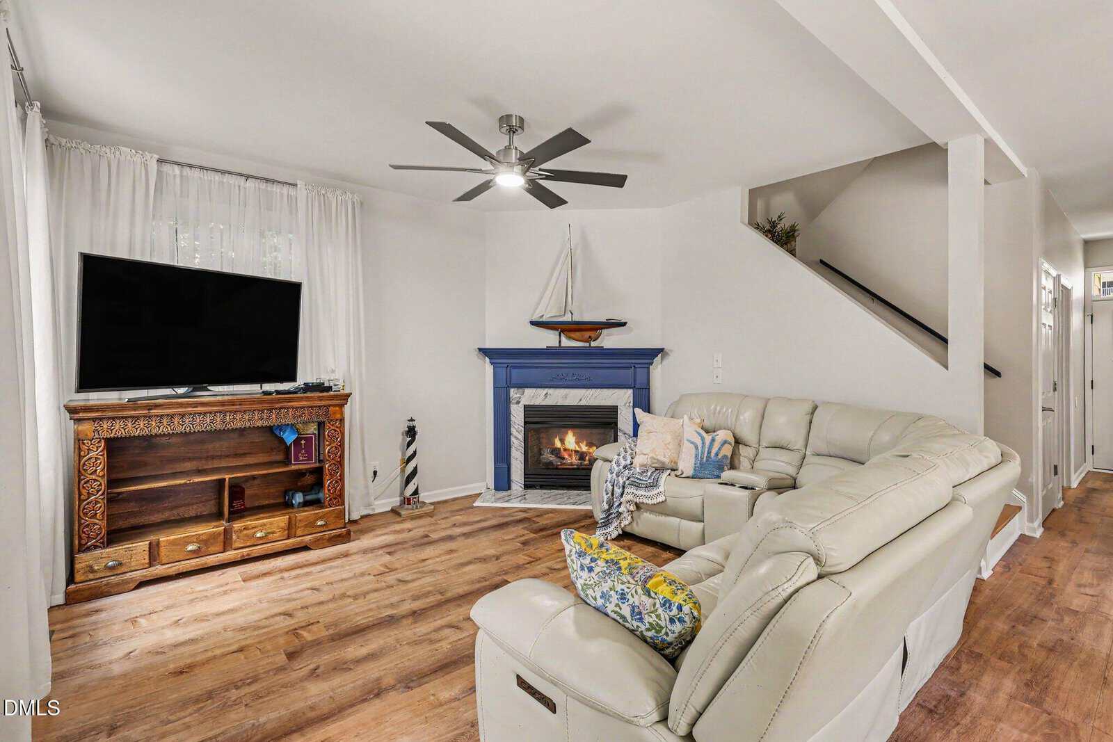 A cozy living room with a white sectional sofa, a TV on a wooden stand, a blue fireplace with a sailboat model above it, wood floors, and a ceiling fan. A staircase is visible in the background.