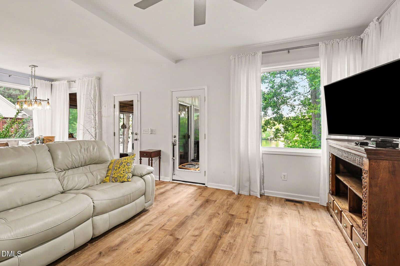 Bright living room with light wood floors, a beige leather sofa, wall-mounted TV on a wooden stand, large windows with white curtains, and a glass door leading outside. Trees visible through the window.