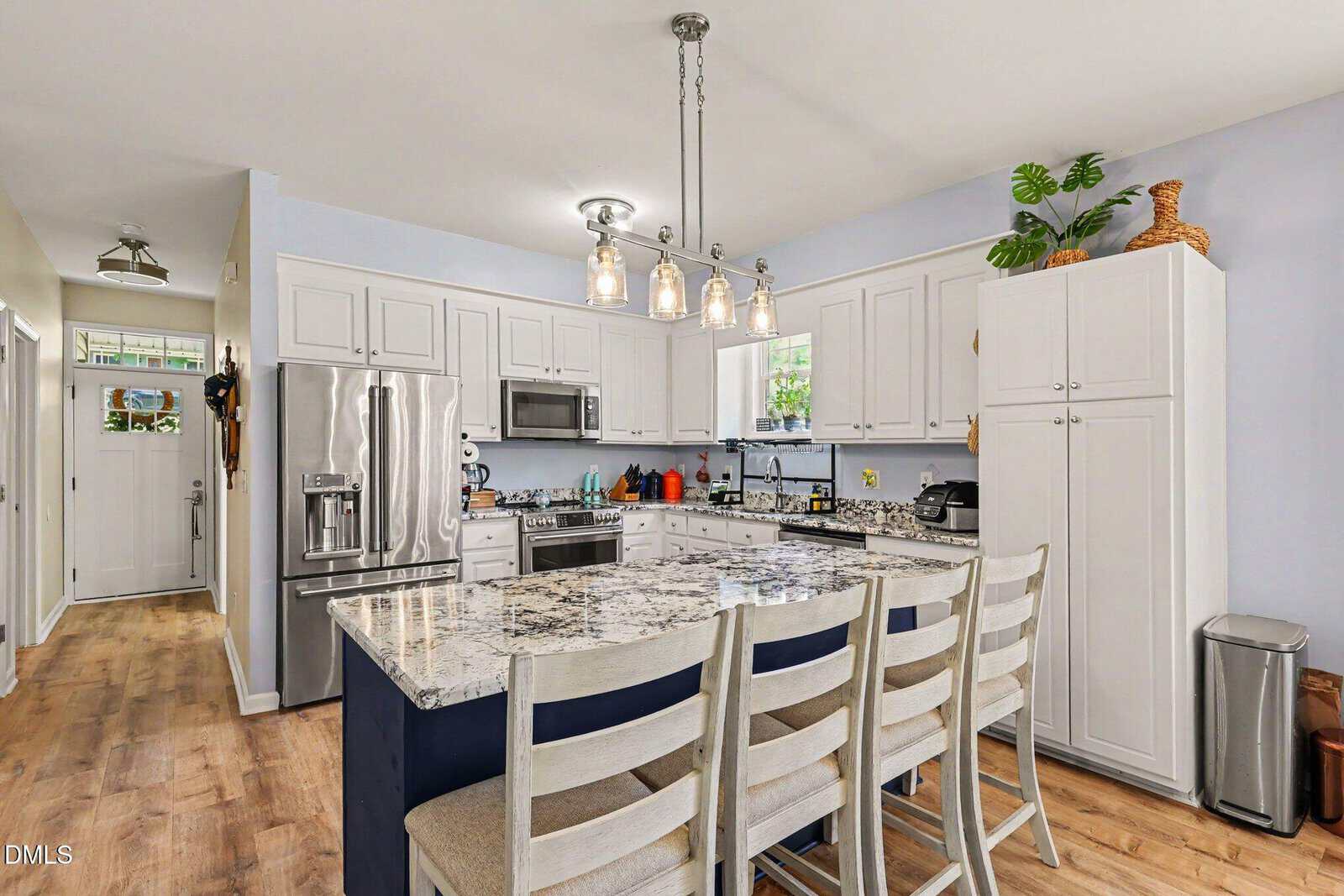 Modern kitchen with white cabinets, stainless steel appliances, marble island with three chairs, light fixture above, wood flooring, and a door leading to a hallway. Potted plant and kitchen essentials add decor.