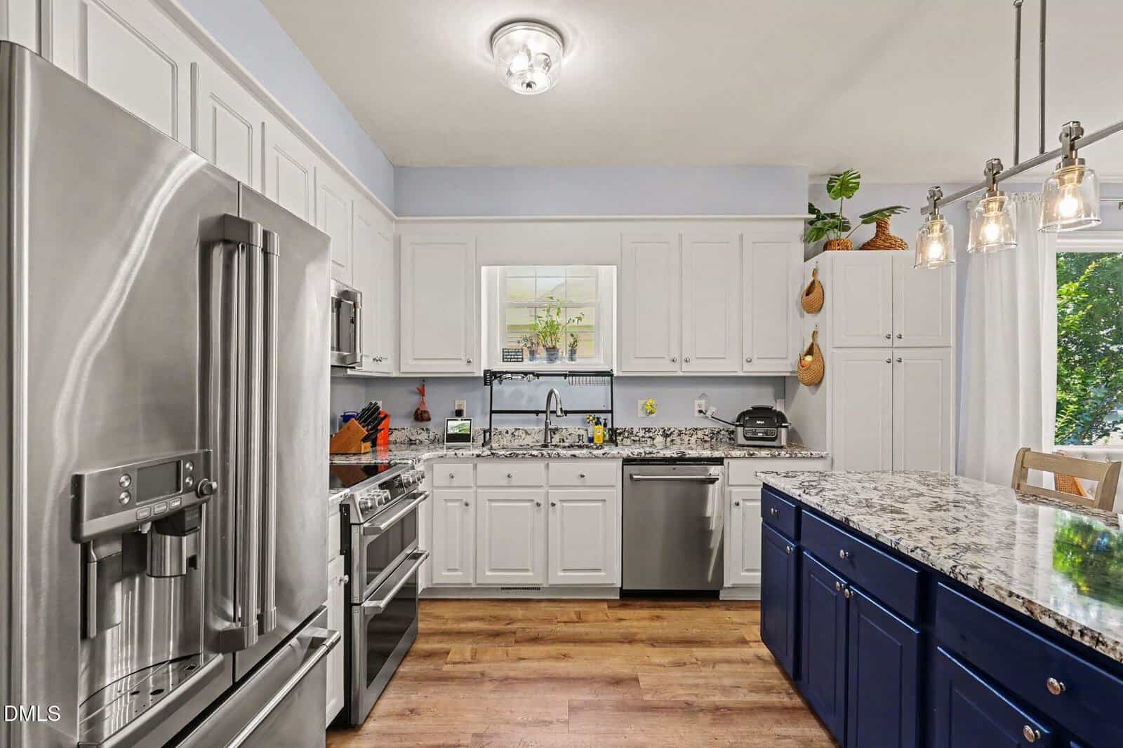 Modern kitchen with white cabinets, stainless steel appliances, blue island, granite countertops, wood flooring, and plants on top of cabinets. Large window lets in natural light, brightening the space.