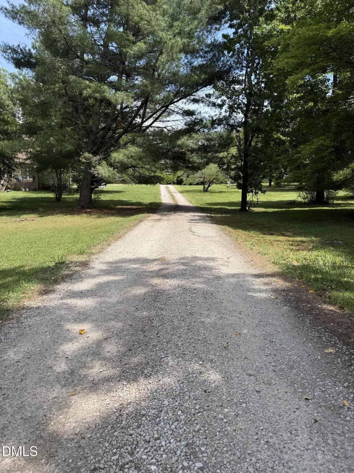 A long gravel driveway stretches through a grassy yard lined with tall green trees, leading toward a house partially visible in the background under a partly sunny sky.