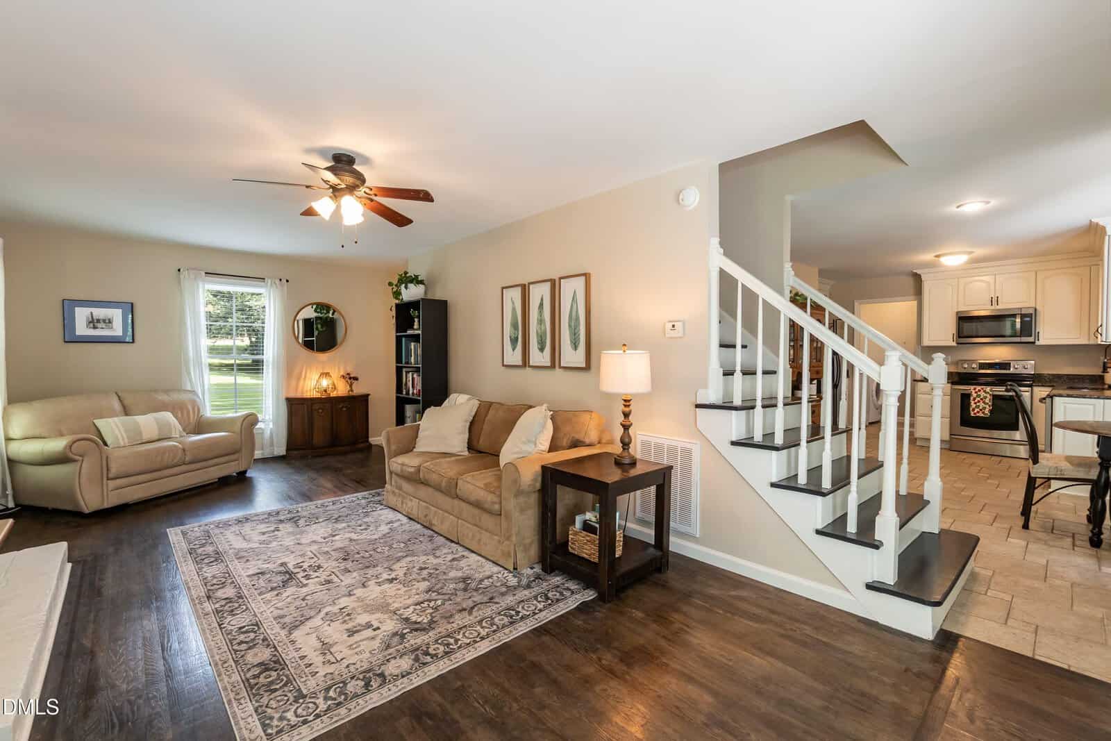 A cozy living room with a beige sofa, armchair, patterned rug, wooden floors, and a staircase leading to an open kitchen with white cabinets and dining area in the background.