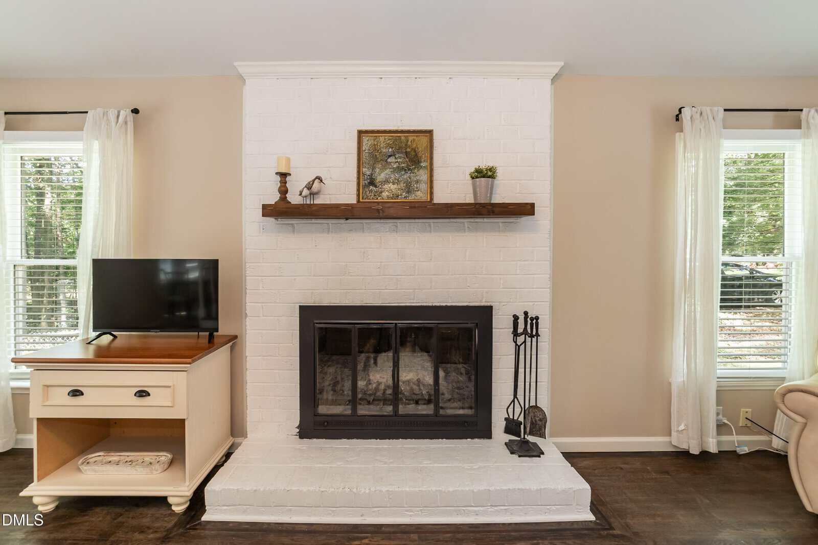 A white brick fireplace with a wood mantel holds decor and art. A black fireplace screen is in front. To the left is a TV on a cream stand; to the right, beige armchair. Two windows with white curtains let in light.