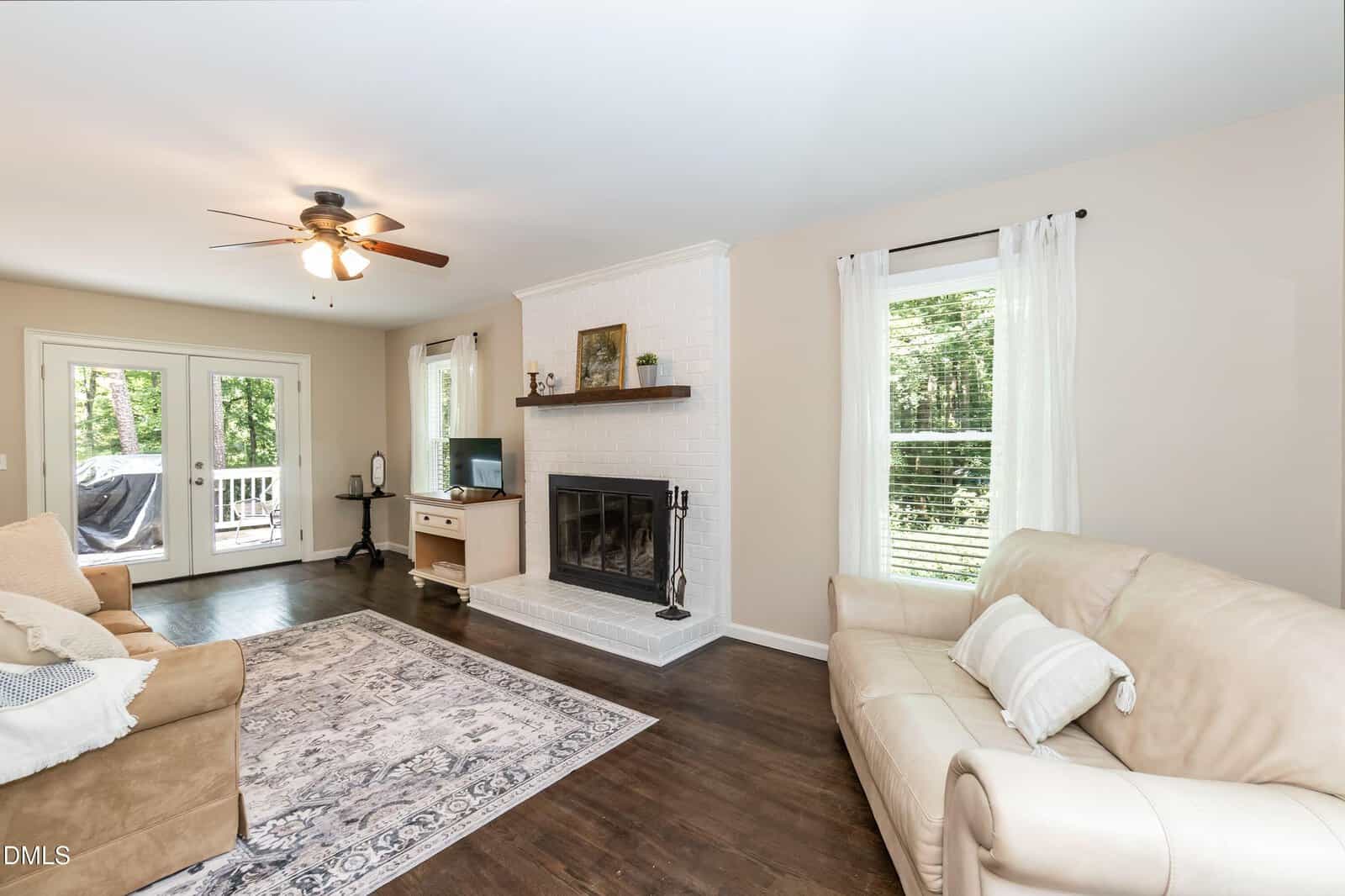 Bright living room with a ceiling fan, beige walls, white fireplace, dark hardwood floor, area rug, cream sofas, TV on a stand, large windows with white curtains, and glass doors leading to a deck with a view of trees.