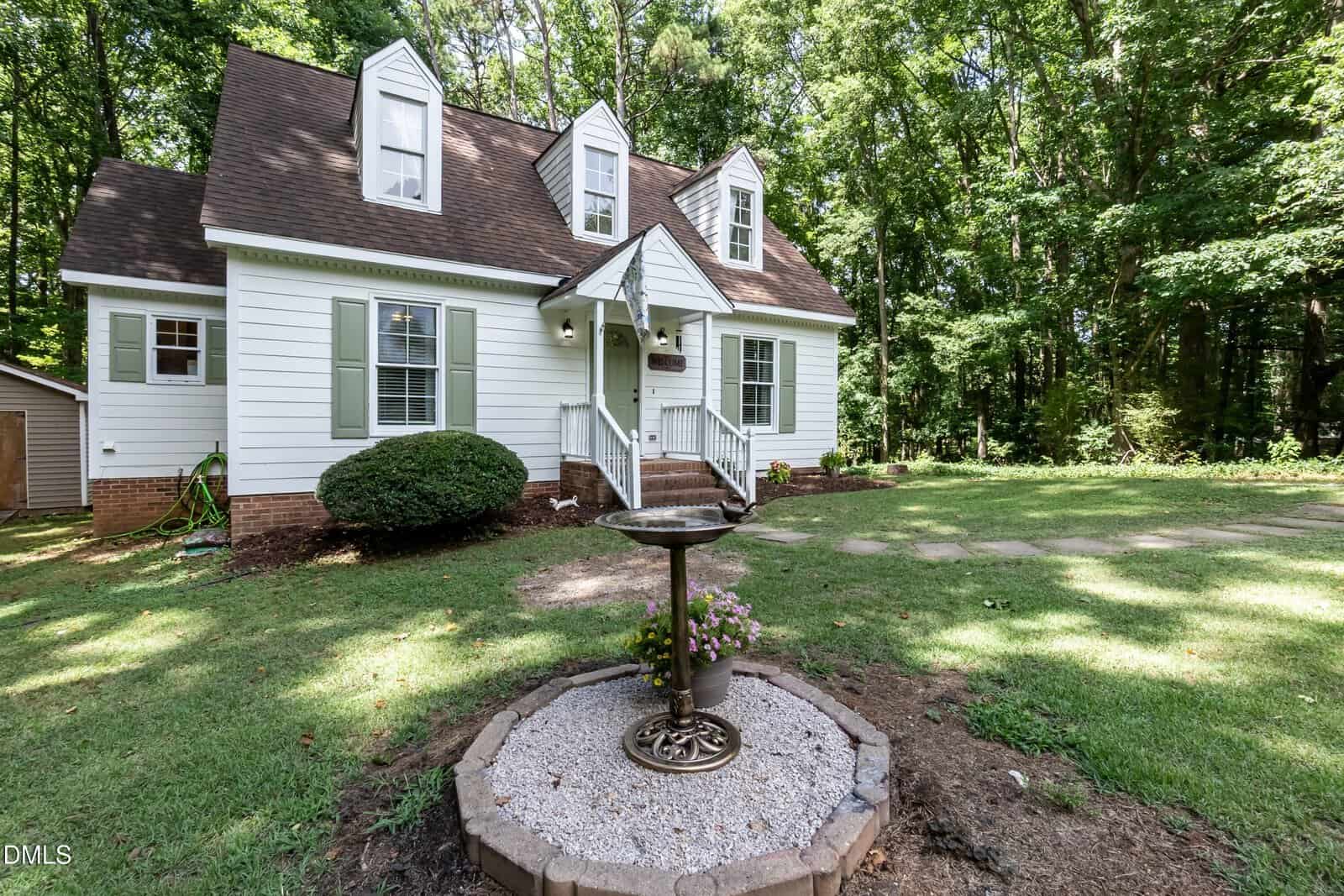 A white two-story house with green shutters and a steep roof sits among tall trees. A birdbath with flowers is in the foreground, surrounded by grass and a shaded yard.