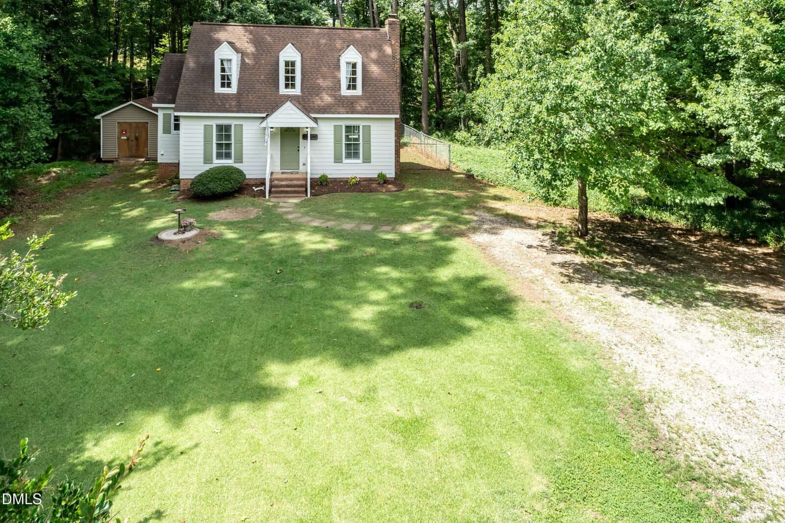A white two-story house with a brown roof and dormer windows sits on a large, grassy yard surrounded by trees, with a gravel driveway on the right and a small wooden shed in the background.