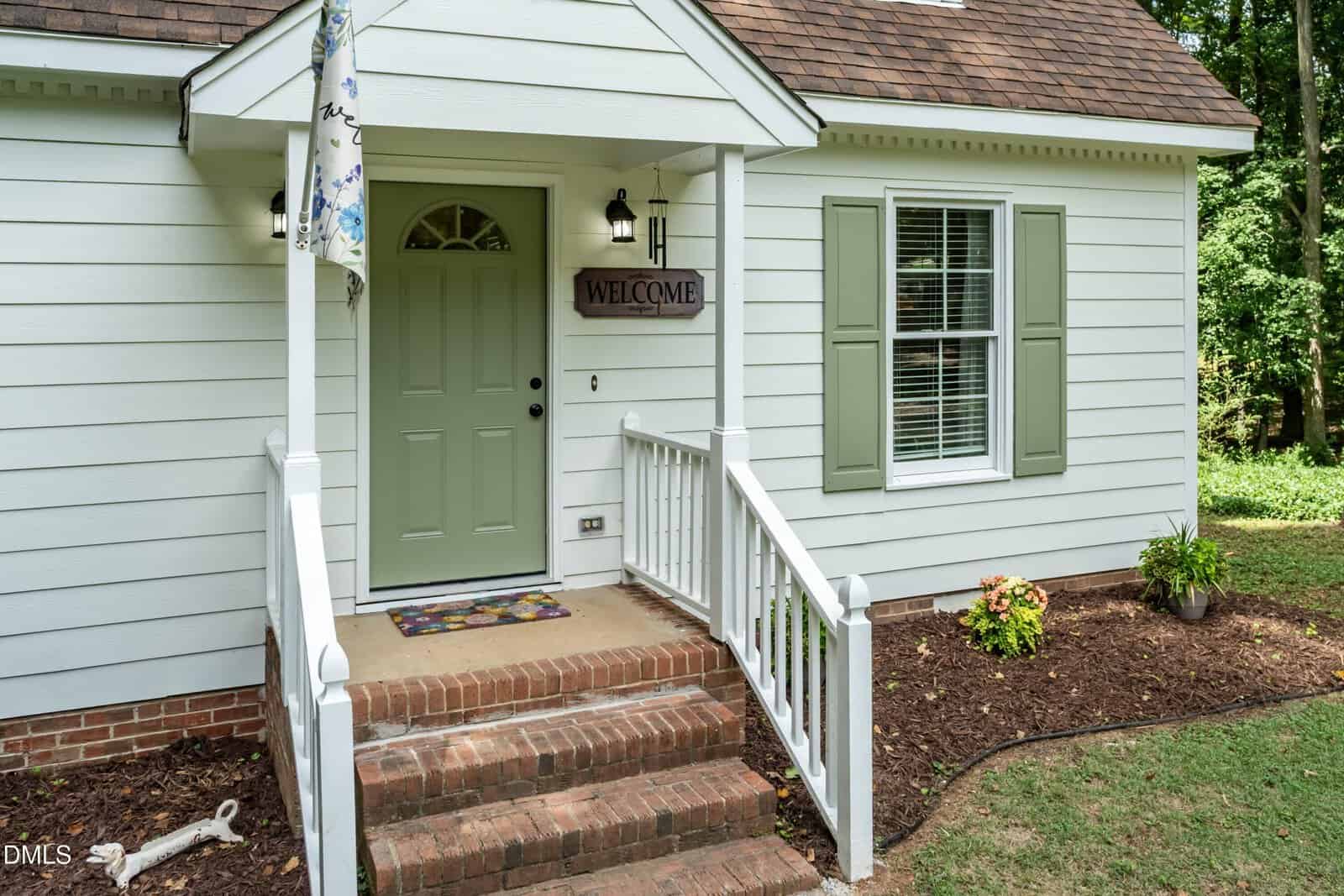 A small house with light green door, white siding, brick steps, and a welcome sign by the entrance. White railings frame the porch, and a doormat and small garden area are visible.