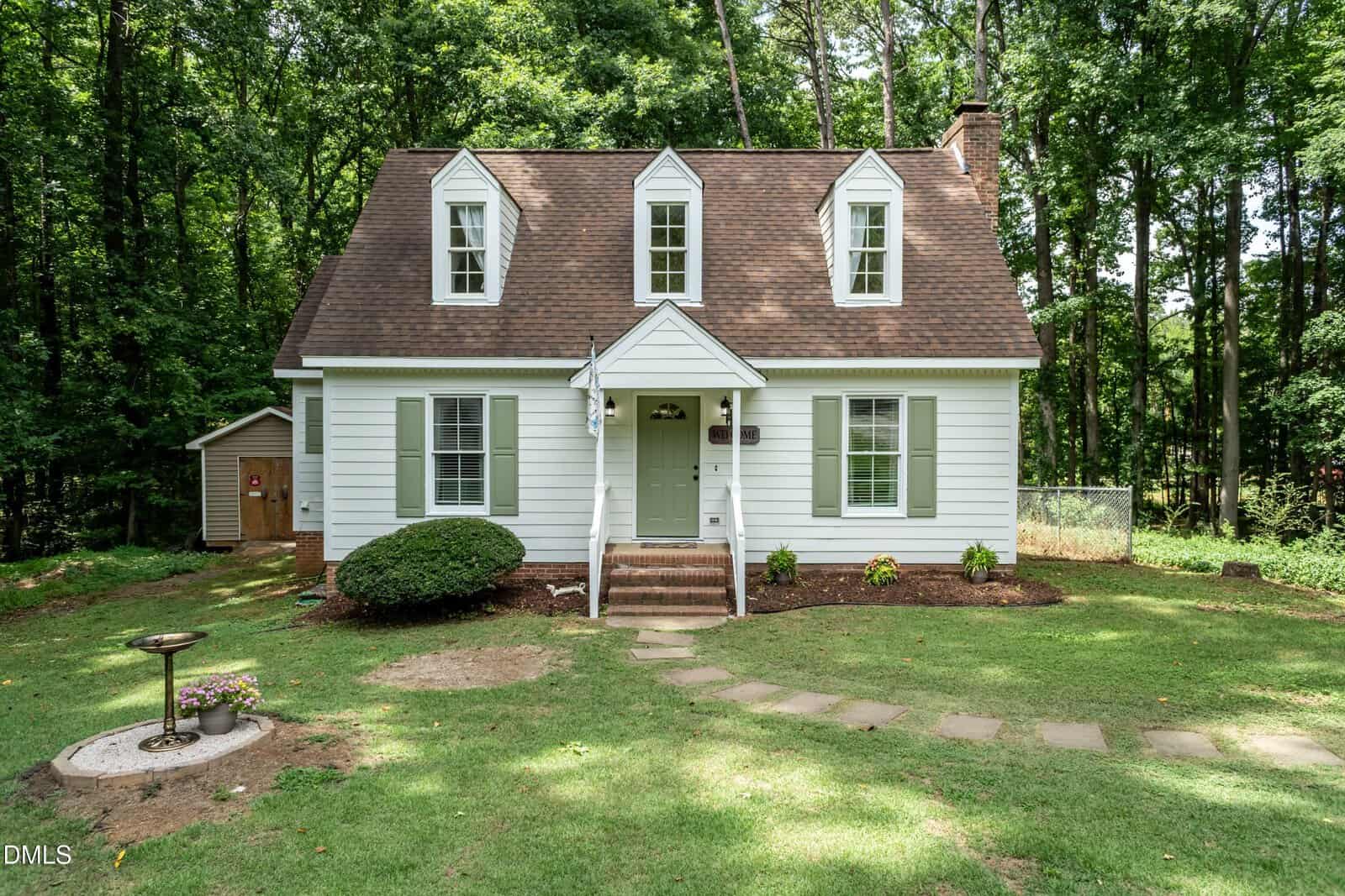 A charming white two-story house with dormer windows and green shutters sits surrounded by grass and trees, featuring a small porch, garden path, birdbath, and shed in the backyard.