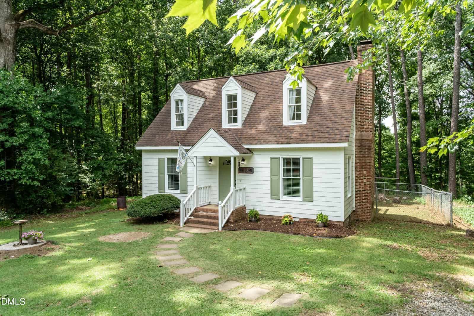 White two-story house with brown roof, dormer windows, and brick chimney. Surrounded by trees, a curved stone path leads to the front steps, with a small garden bed and a fenced yard on one side.