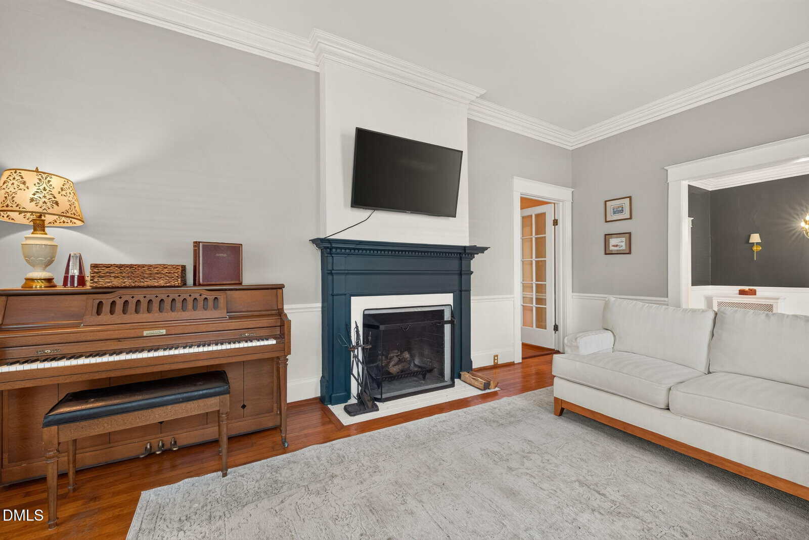 Living room with a piano, fireplace with a mounted TV, white couch, wooden floors, gray walls, and a floor lamp. Doorways lead to other rooms, and framed art is hung on the wall.