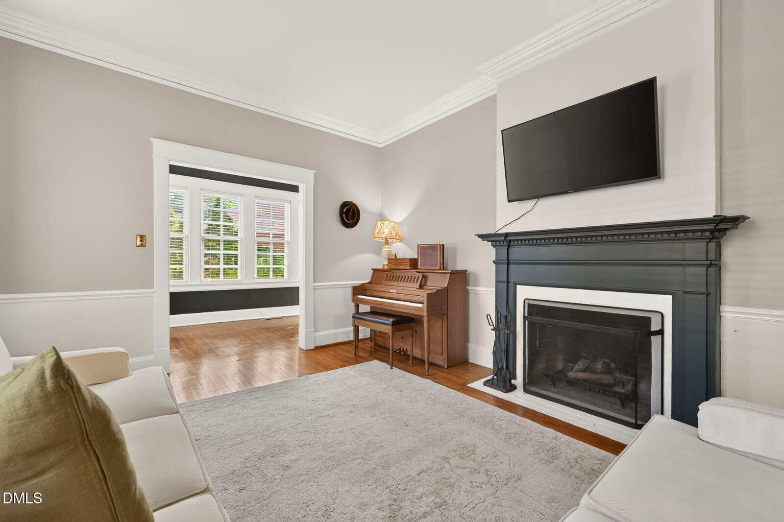 Bright living room with a white sofa, beige rug, black fireplace, wall-mounted TV, piano, lamp, and large window with white shutters. Light wood flooring and neutral walls create an inviting atmosphere.