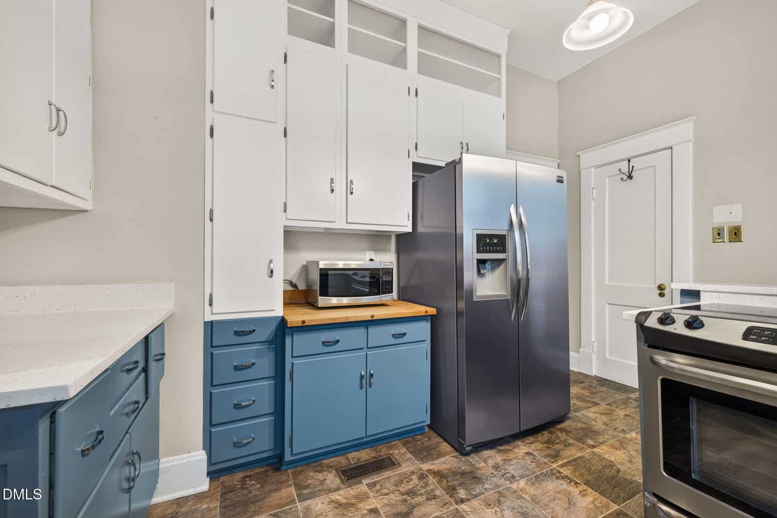 A kitchen with blue lower cabinets, white upper cabinets, a stainless steel refrigerator, a stove, a microwave on a wooden counter, and tile flooring. The walls are light gray with white trim and doors.