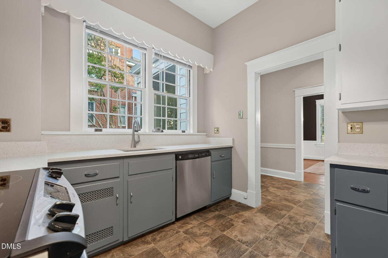 Bright kitchen with gray cabinets, stainless steel dishwasher, stove, and a large window above the sink. The floor is tiled, and there is an open doorway leading to another room.