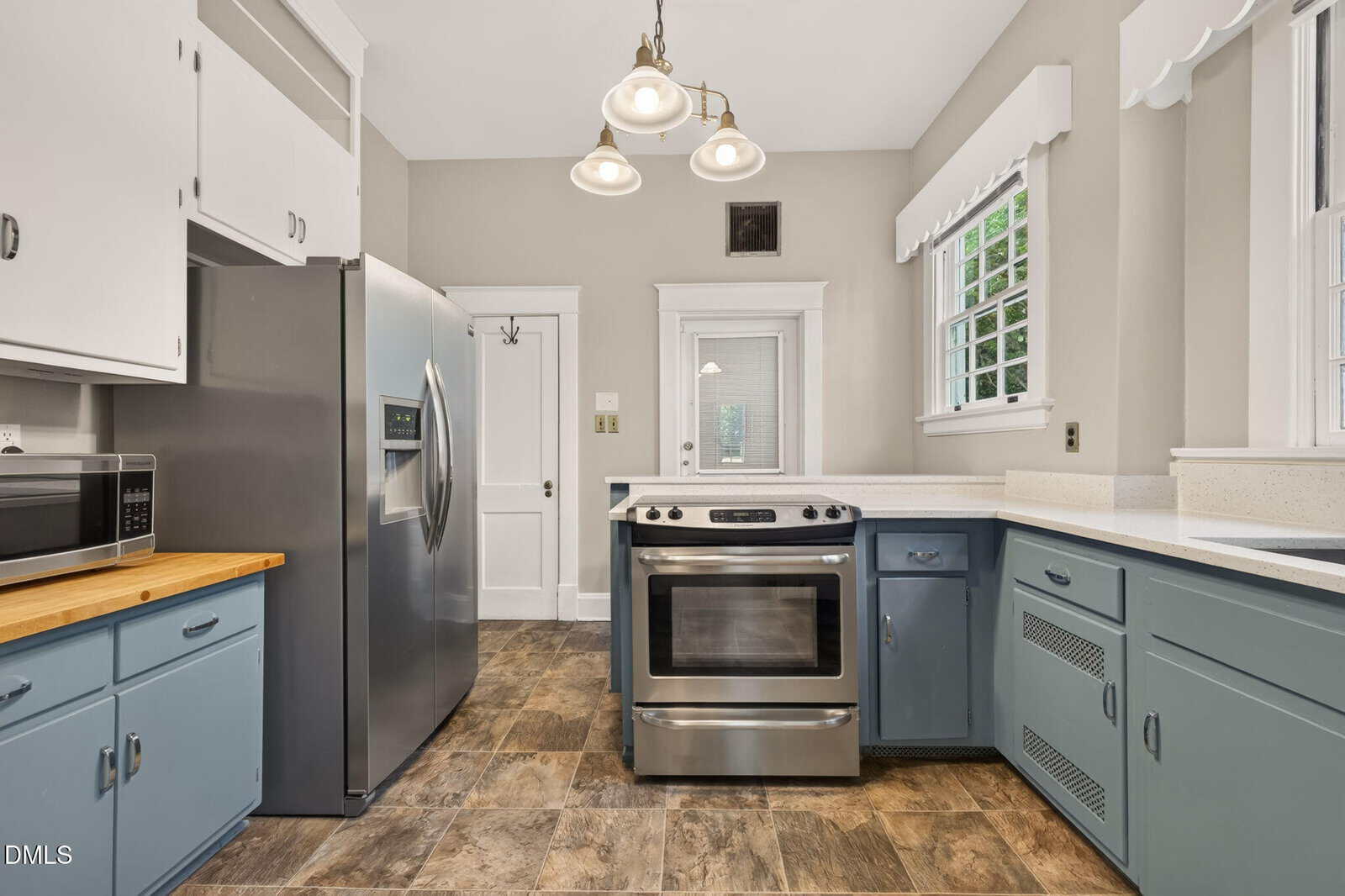 A kitchen with blue lower cabinets, white upper cabinets, stainless steel appliances including a refrigerator and oven, neutral walls, three pendant lights, and windows with white trim and valances.