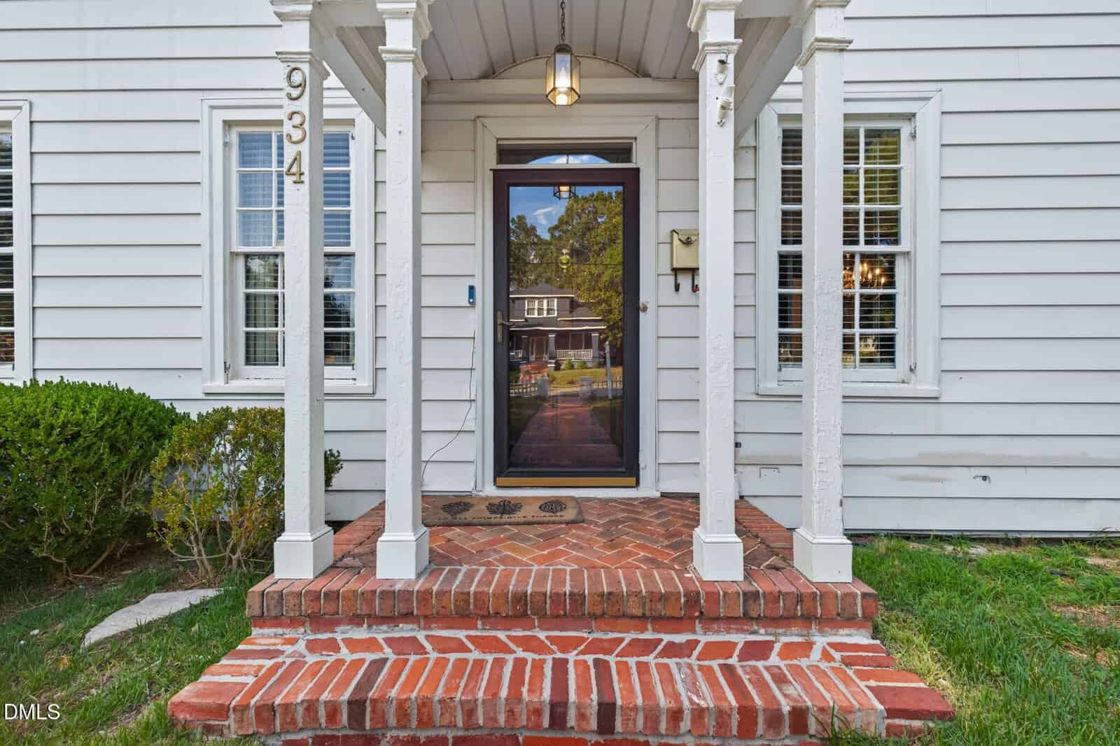 A small brick porch with white pillars leads to a glass front door of a white house with siding. The house number 934 is displayed beside the door. Bushes and grass surround the porch.