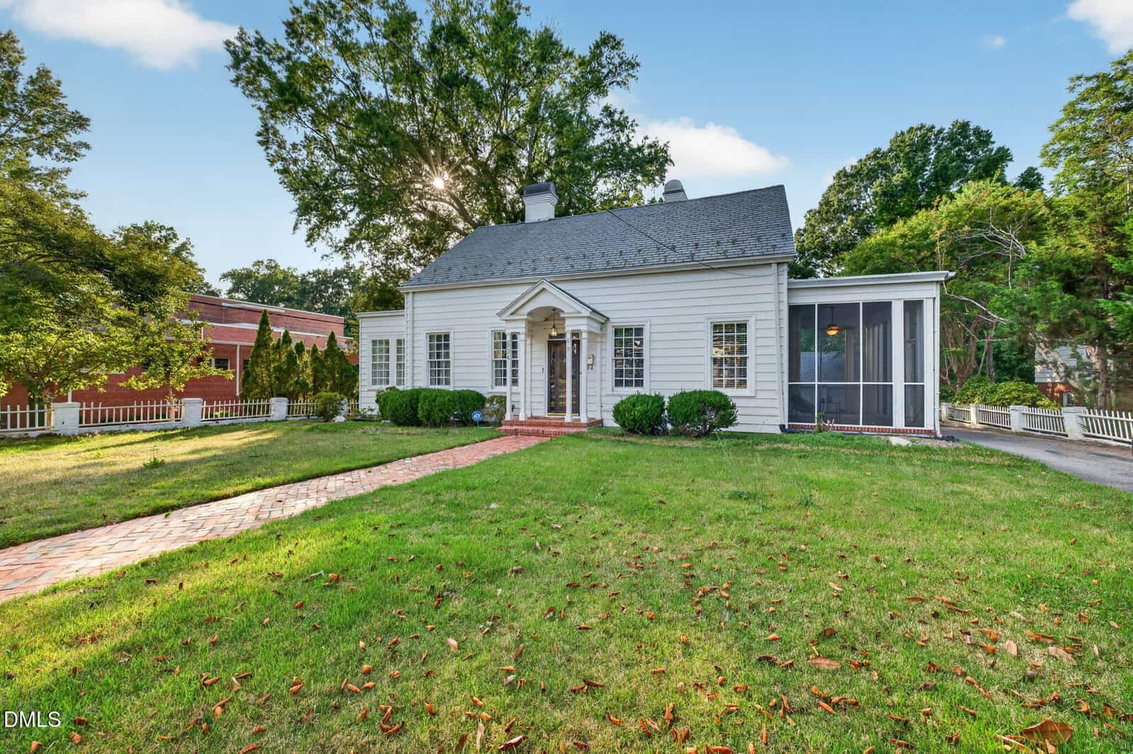 A white, single-story house with a front porch and columns, a brick walkway, green lawn, and screened side porch, surrounded by trees and a white fence under a blue sky.