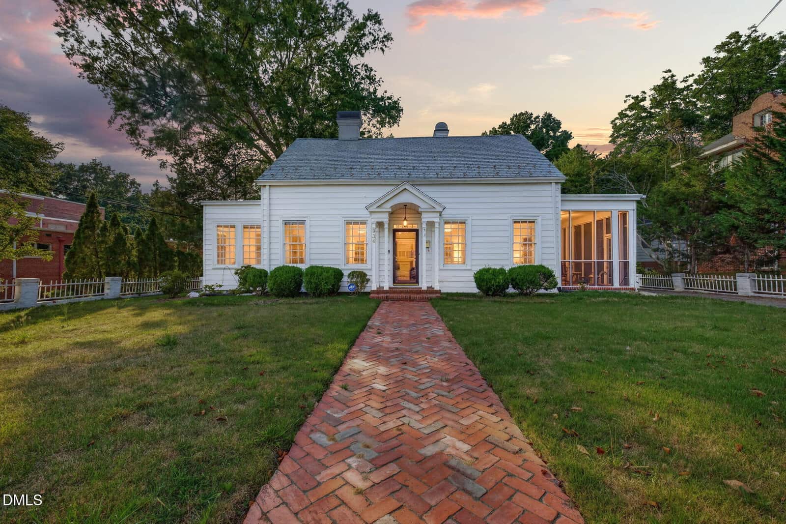 A charming white house at 934 S Chestnut Street, Henderson, with glowing windows sits at the end of a red brick walkway, surrounded by a green lawn, bushes, and trees at dusk under a pink and blue sky.