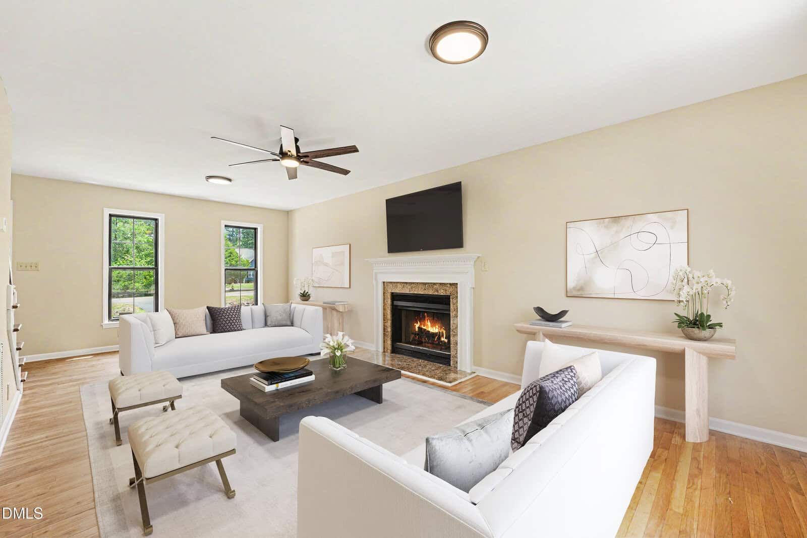 Bright living room with light walls, hardwood floors, two white sofas, a coffee table, a fireplace with a TV above it, two benches, wall art, windows, and a ceiling fan.