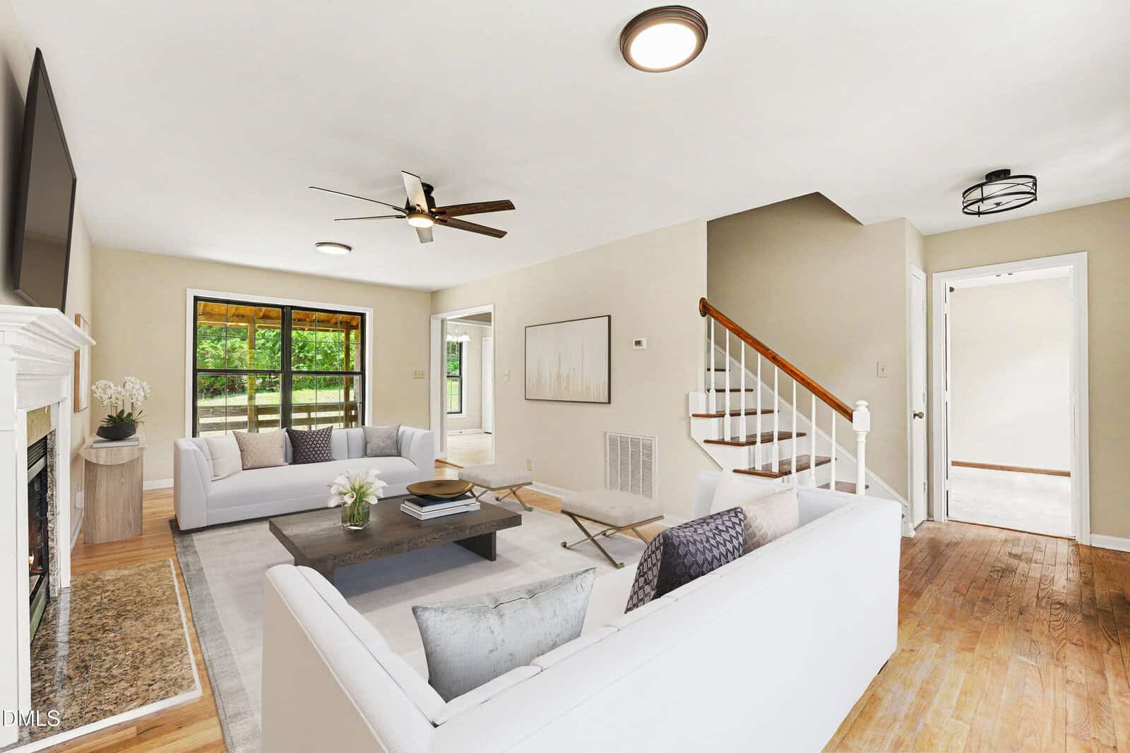 Bright living room with two white sofas, a coffee table, a fireplace, wooden floors, a ceiling fan, and stairs leading up. Large windows let in natural light, and neutral decor creates a welcoming atmosphere.