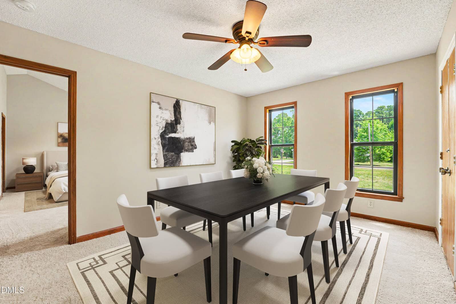 A dining room with a black table, eight white chairs, a light-colored rug, a ceiling fan, two large windows, a modern art piece, and an open doorway leading to a bedroom.
