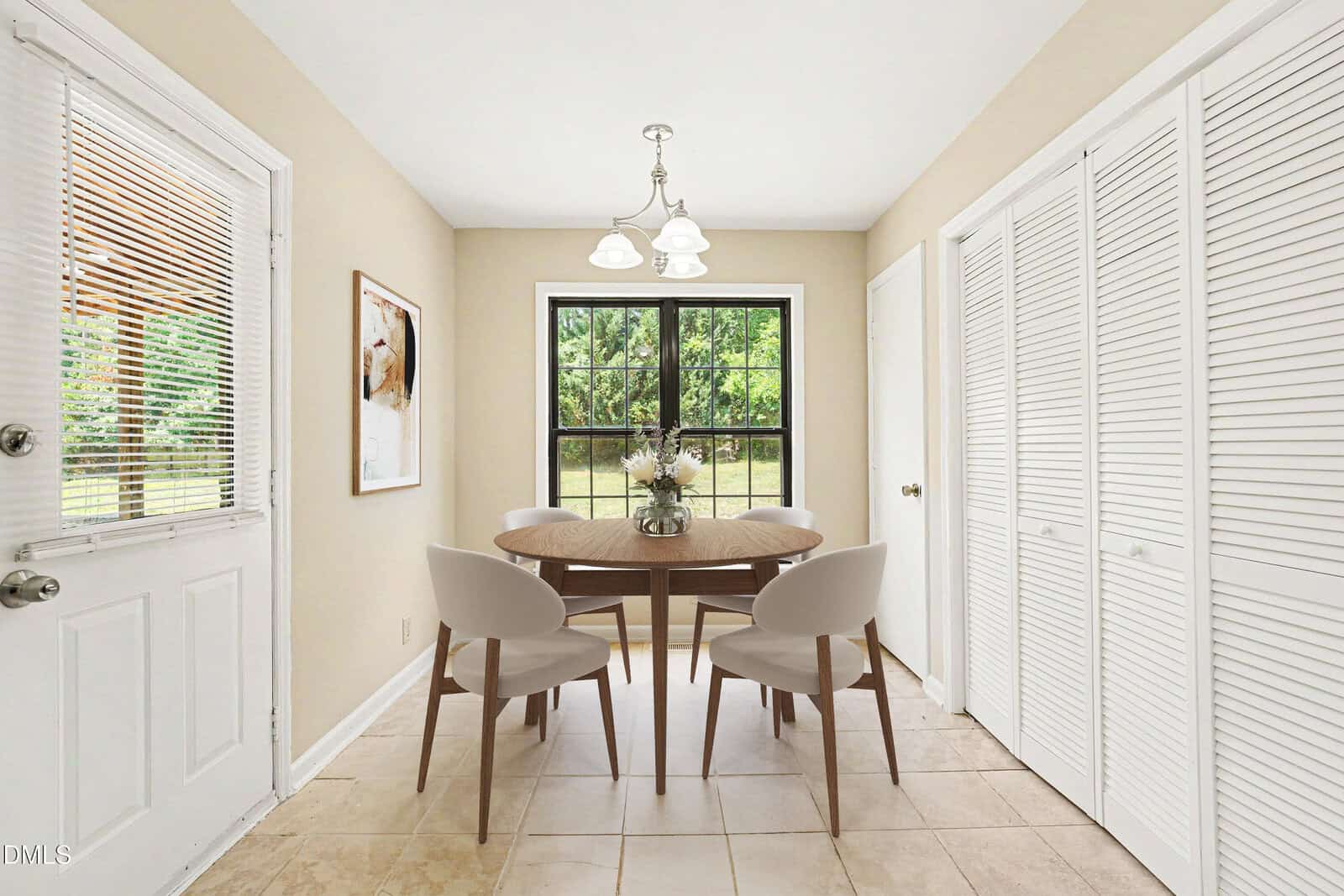 A bright dining area with a round wooden table, four beige chairs, a hanging light fixture, a window overlooking greenery, beige walls, tile flooring, and closets with white louvered doors.
