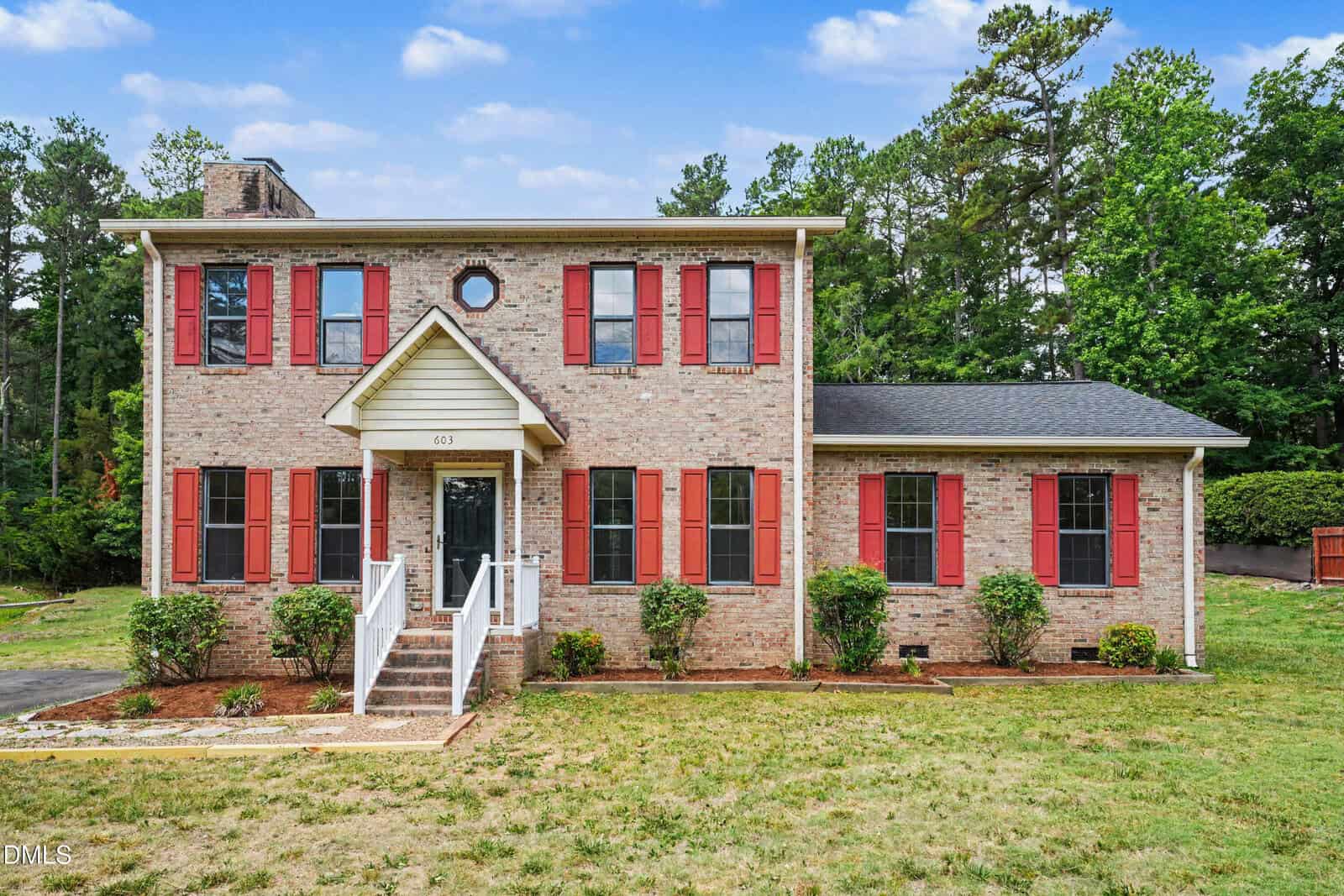Charming two-story brick house at 603 20th Street in Butner, featuring red shutters and a covered front entrance with white railings. The grassy yard is dotted with small shrubs, and mature trees stand under a partly cloudy sky.
