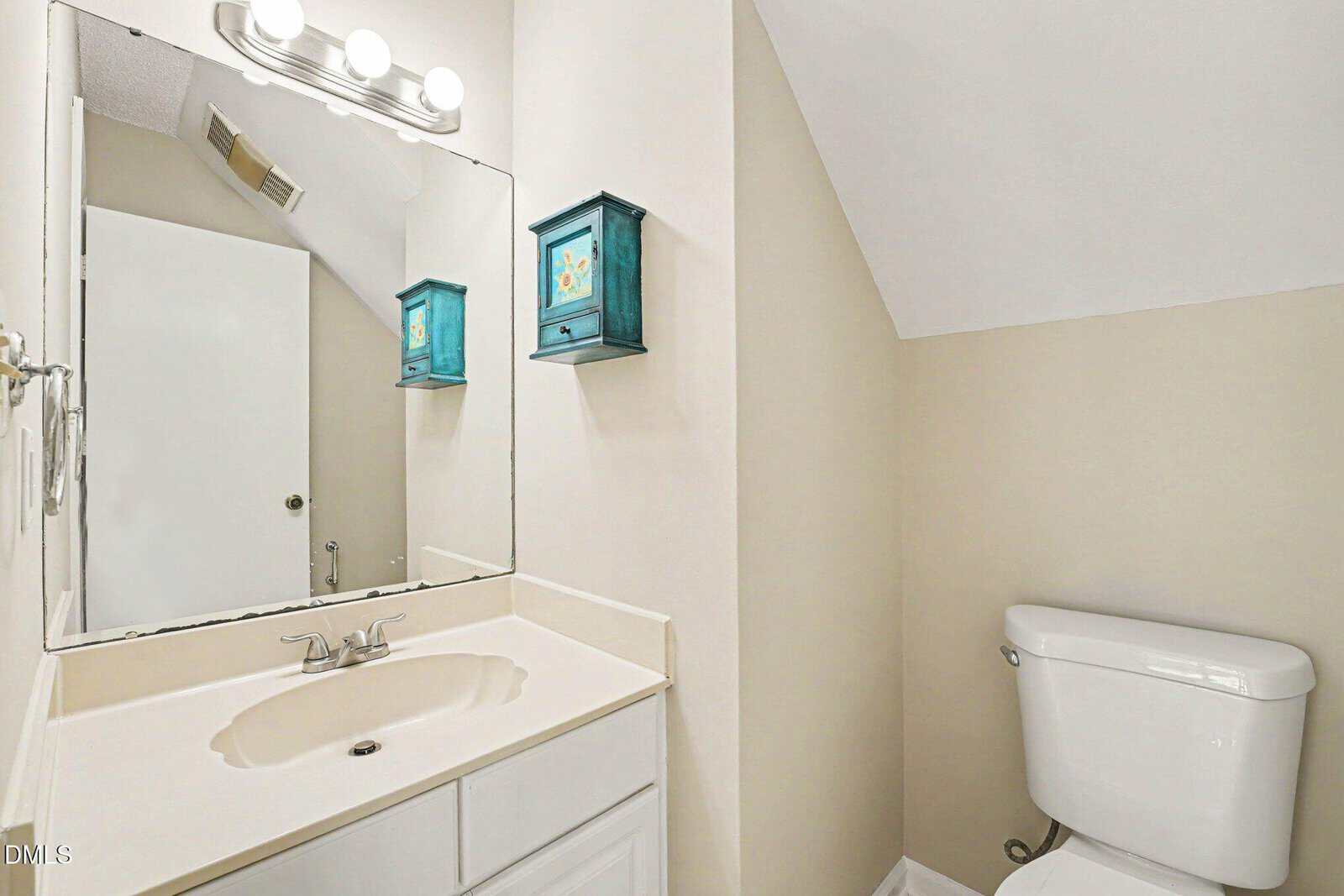 A bathroom with a white sink and vanity, a wall mirror with lights above, a white toilet, and blue wall-mounted cabinets on a beige wall under a sloped ceiling.
