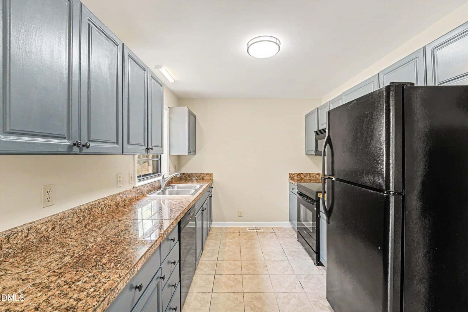 Narrow kitchen with blue cabinets, black appliances, granite countertops, double sink under a window, and beige tiled floor. Walls are a light neutral color, and ceiling light fixtures are present.