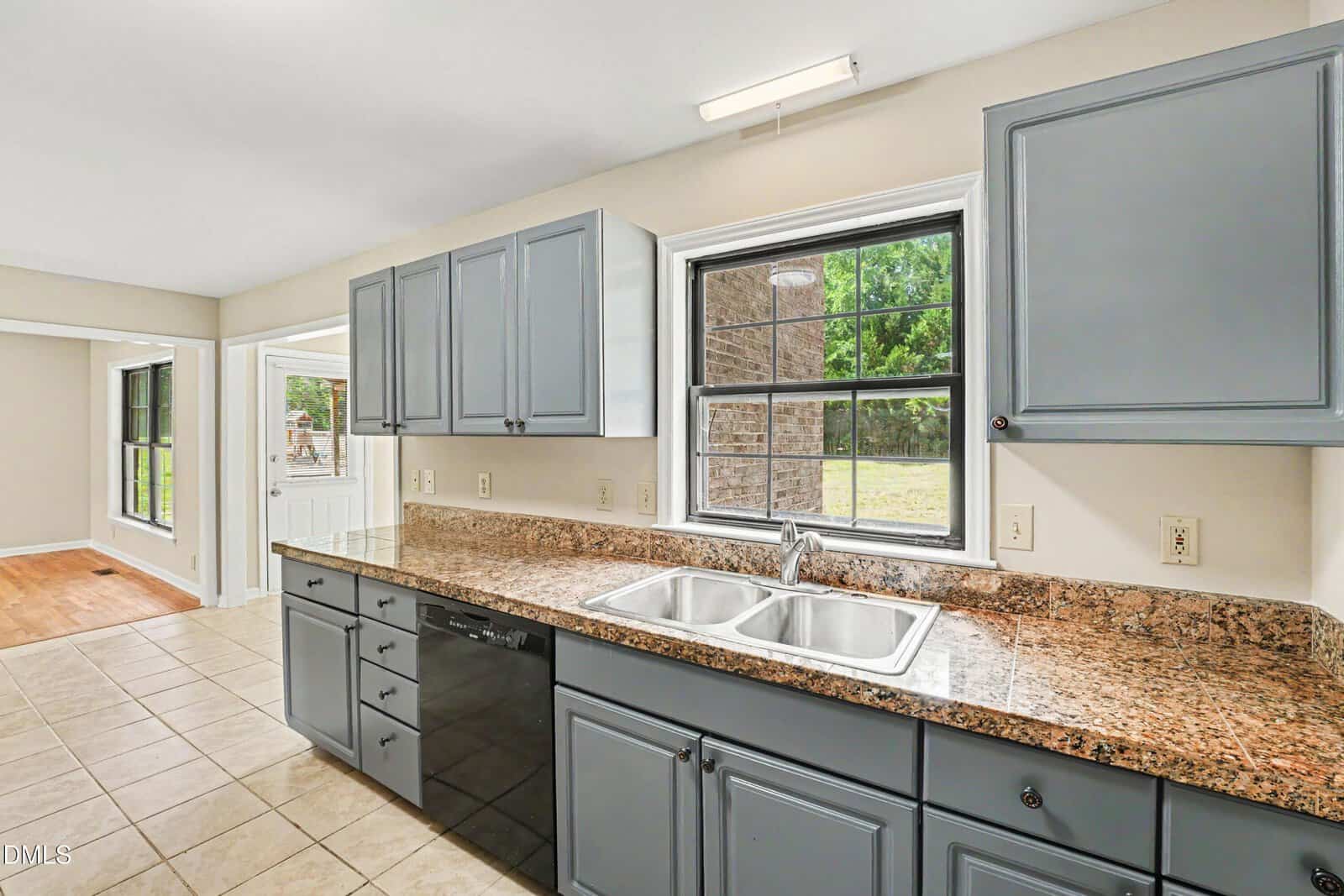 Modern kitchen with gray cabinets, a double stainless steel sink, granite countertops, black dishwasher, tile flooring, and a window above the sink overlooking a green backyard. Adjoining room with wood flooring is visible.