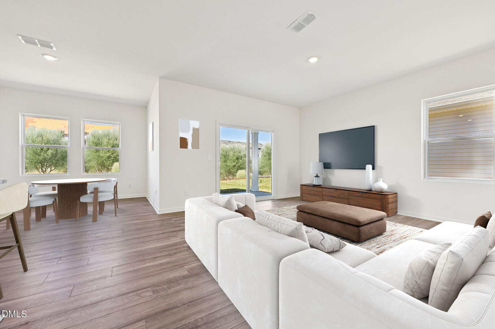 Bright living room with white walls, wood floors, a white sectional sofa, a brown ottoman, a large TV on a wooden console, dining table by windows, and sliding doors opening to a green backyard.
