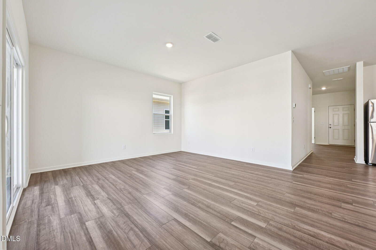Empty, unfurnished room with light-colored walls, wood-style flooring, one window, and sliding glass doors. The space is open with visible kitchen appliances and hallway leading to other rooms.