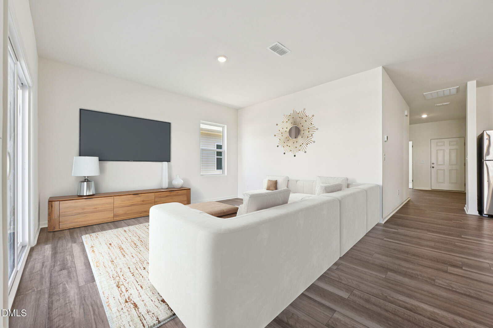 Modern living room with a large white sectional sofa, wood flooring, TV on a wooden console, decorative mirror on the wall, window, and open view to hallway and kitchen. Neutral colors create a bright, airy atmosphere.