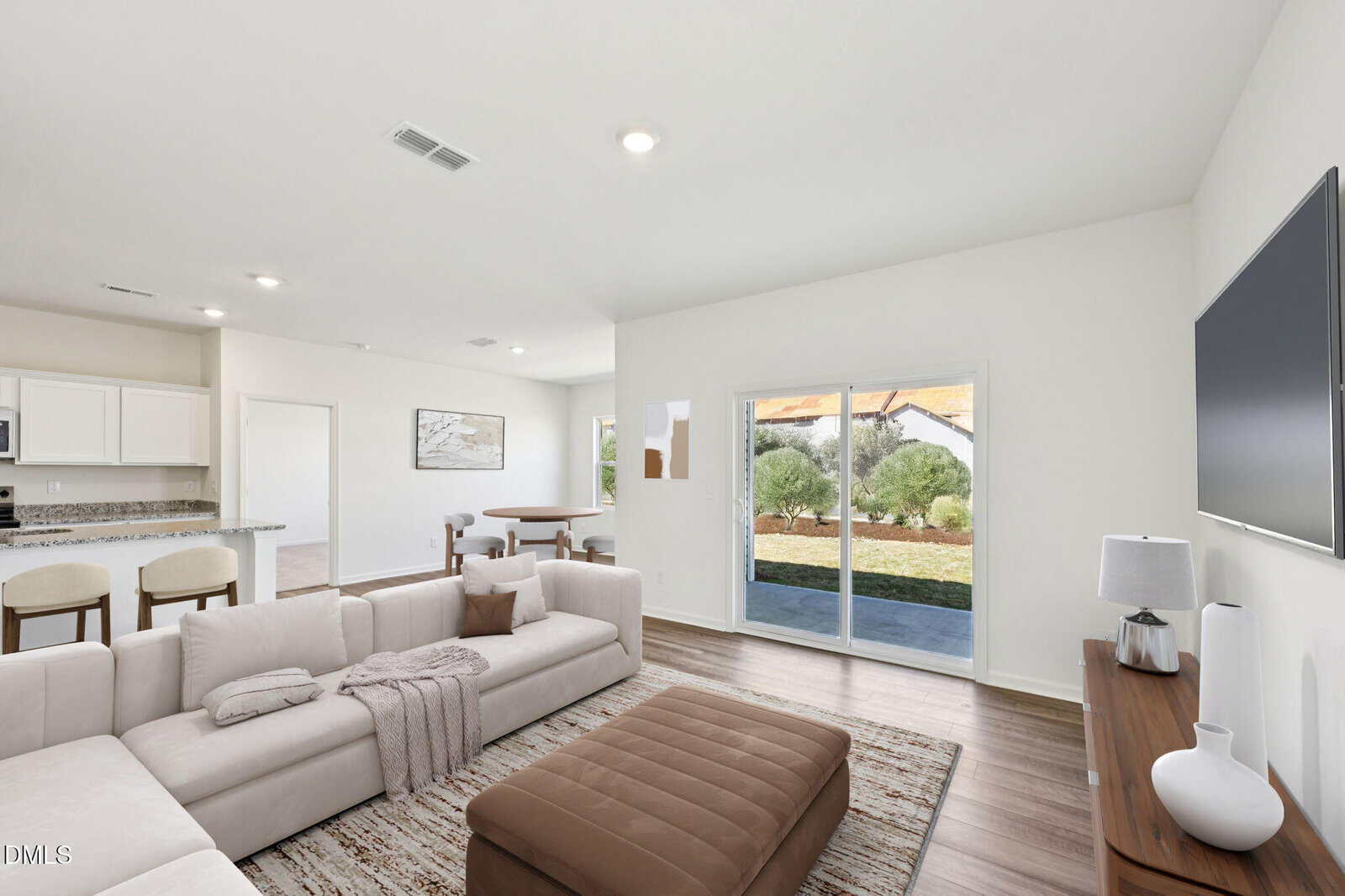 A modern living room with white walls, light wood flooring, a beige sectional sofa, ottoman, TV, and sliding glass doors opening to a backyard with green grass and bushes. A dining area and kitchen are visible in the background.