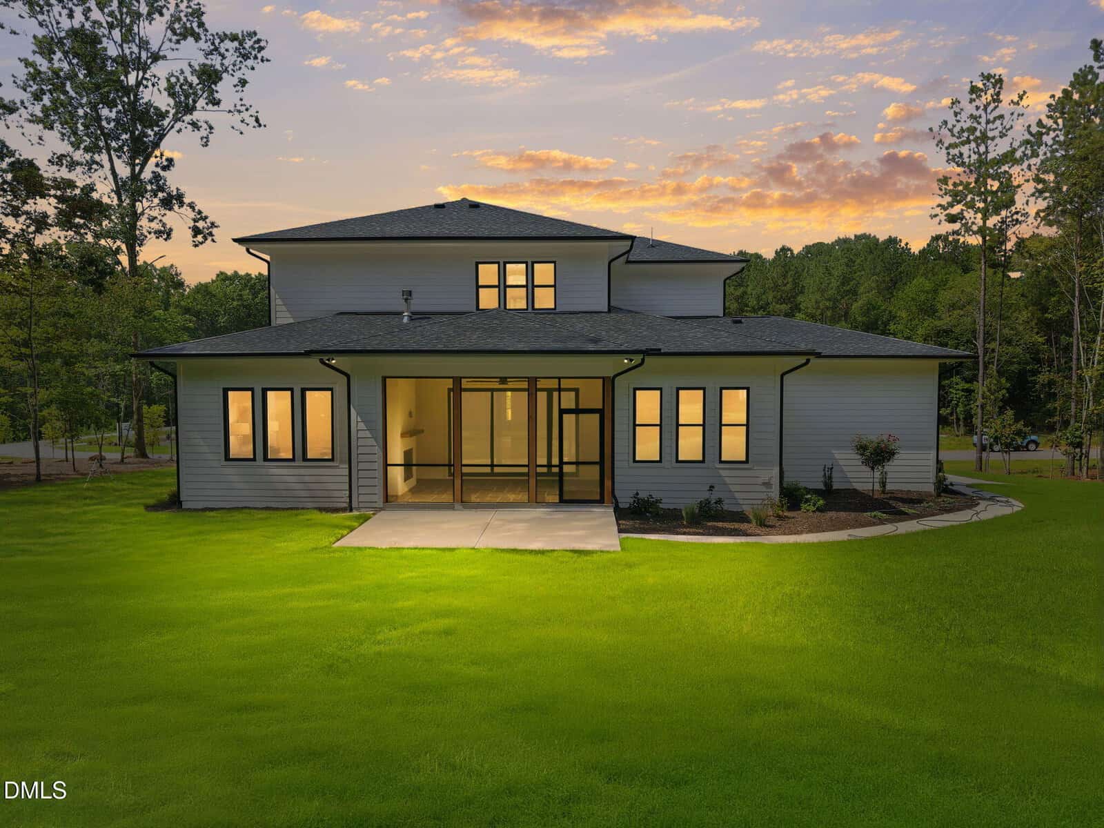 Modern two-story house with white siding and large windows, illuminated at dusk. The home has a covered entryway and sits on a spacious, well-maintained lawn, surrounded by trees under a colorful sunset sky.
