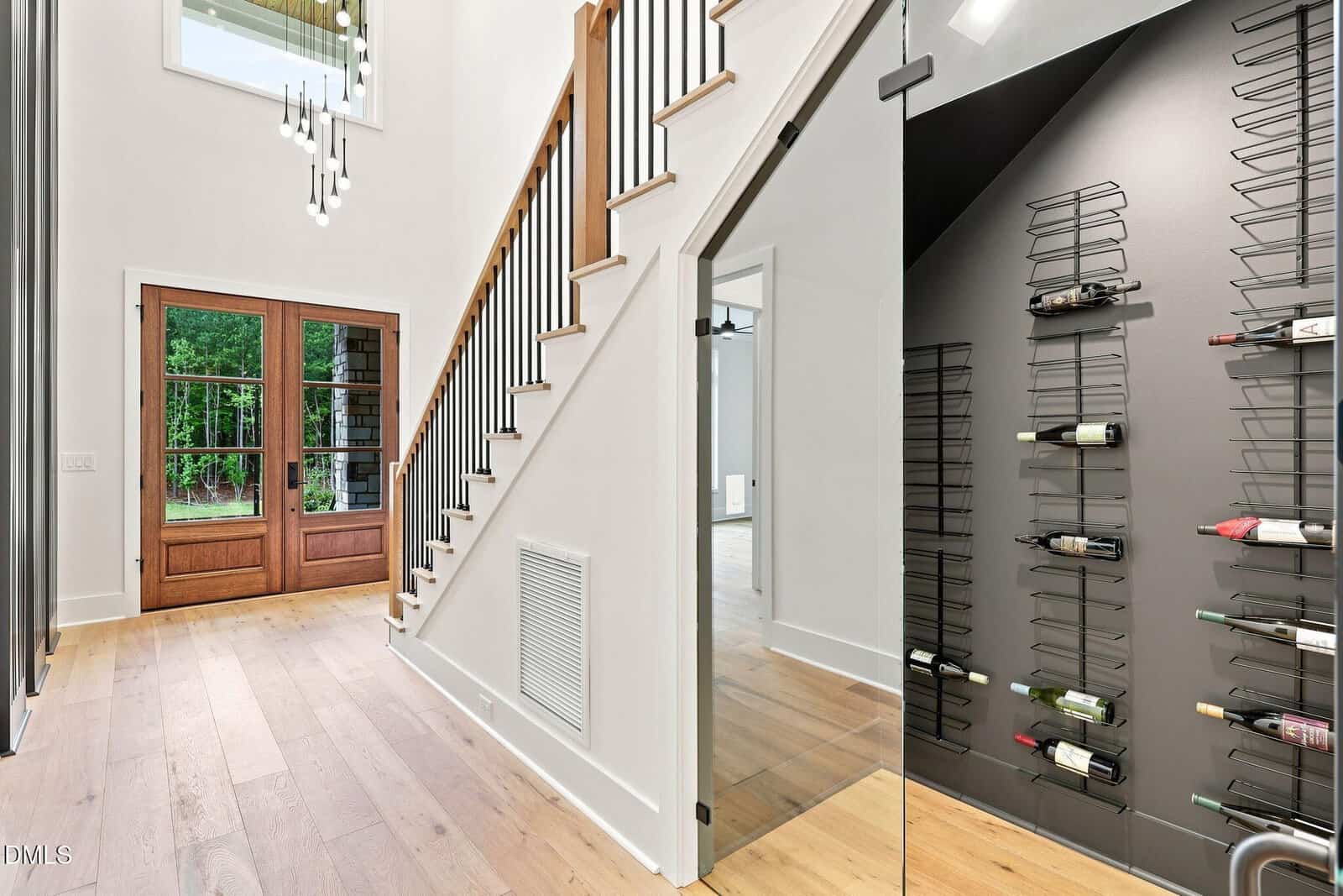 A modern home entryway with wood floors, a staircase with black railings, glass wine storage under the stairs, a tall chandelier, and double wooden doors opening to a green outdoor area.