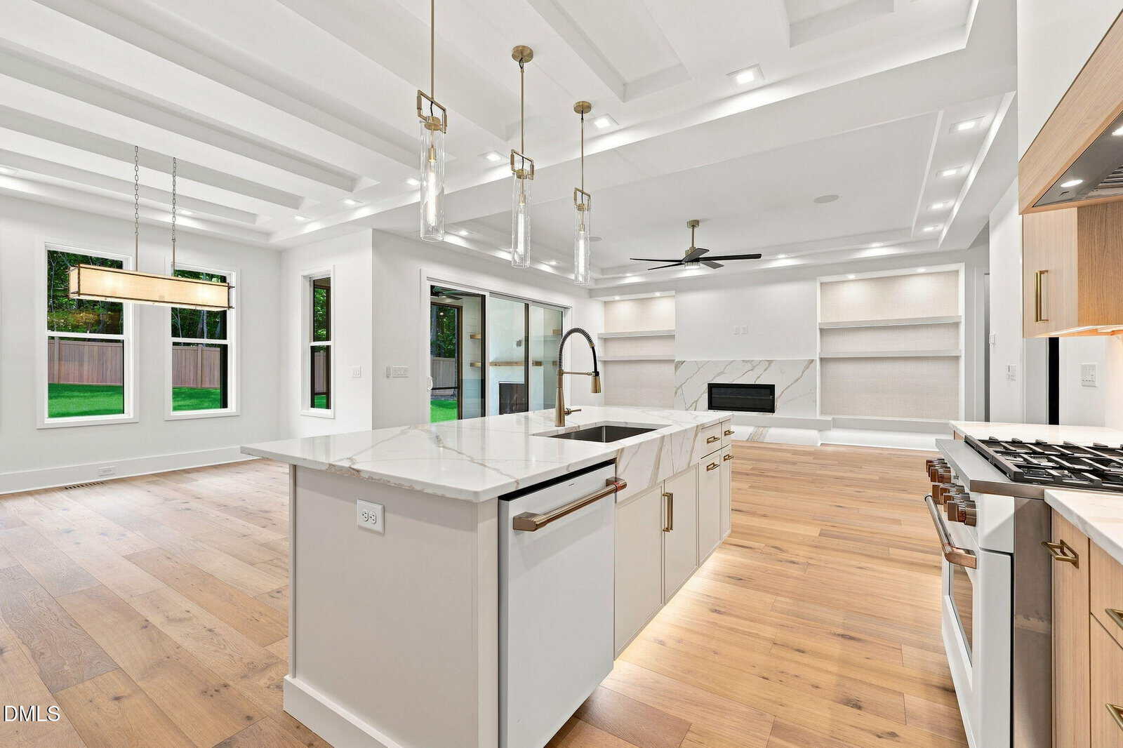 Modern, open-concept kitchen and living area with wood floors, white cabinets, marble countertops, pendant lighting, built-in shelves, fireplace, large windows, and sliding glass doors leading to a green backyard.