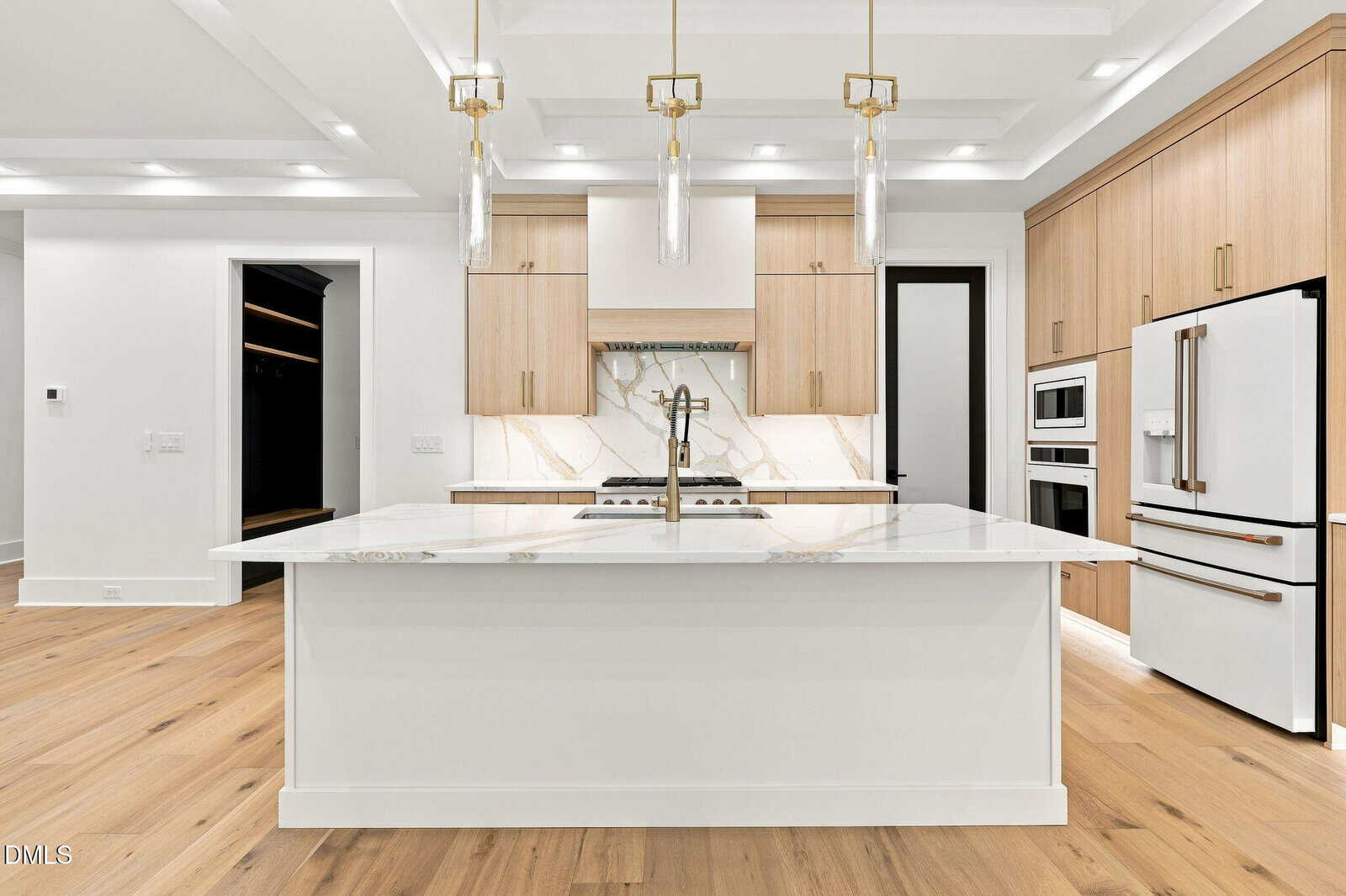 Modern kitchen with light wood cabinets, white marble island and backsplash, three pendant lights, double wall ovens, and a built-in fridge. Minimalist design with light wood flooring and recessed ceiling details.