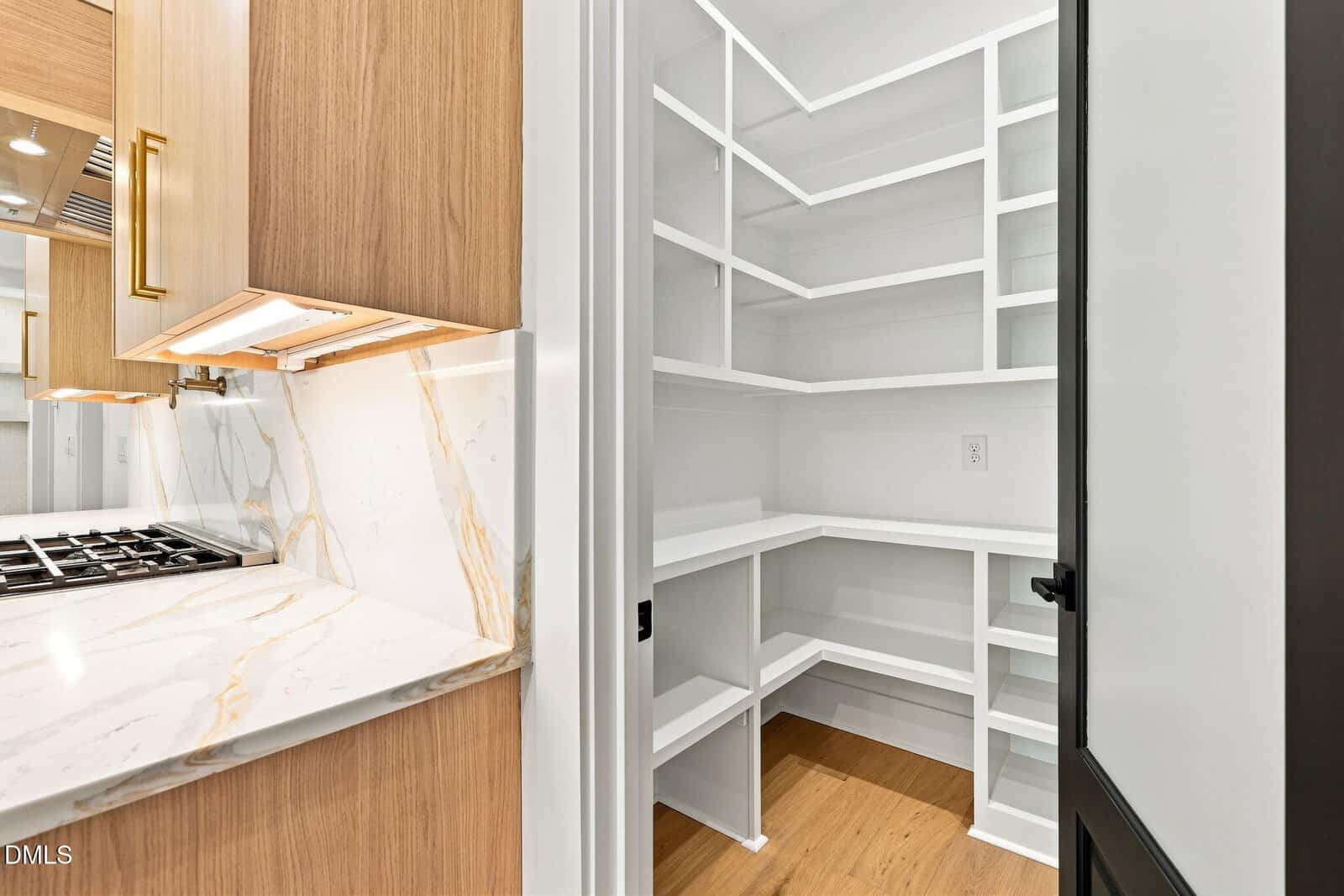A clean, empty walk-in pantry with white shelves is next to a kitchen with wooden cabinets, gold hardware, and a marble countertop.