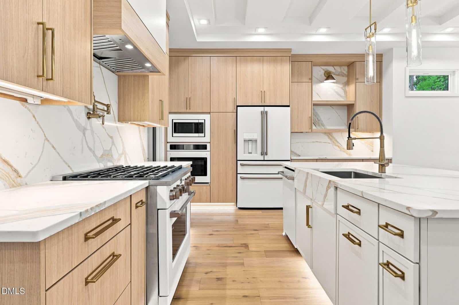 Modern kitchen with light wood cabinets, white marble countertops and backsplash, stainless steel appliances, double wall oven, gas stove, built-in refrigerator, large island with sink, pendant lights, and wood flooring.