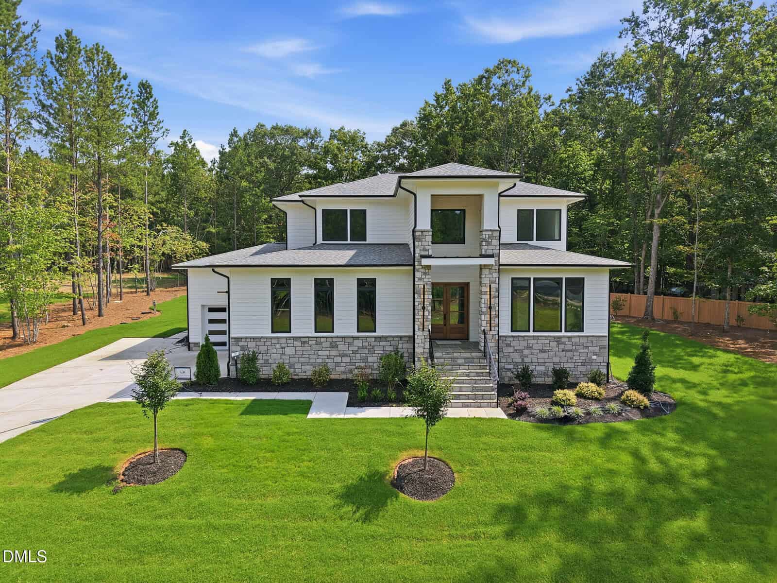 Two-story modern house with stone and white siding, large windows, a covered entryway, and landscaped front yard at 20 Cherry Bark Drive. Surrounded by green grass, small trees, and wooded areas under a blue sky with light clouds in serene Youngsville.